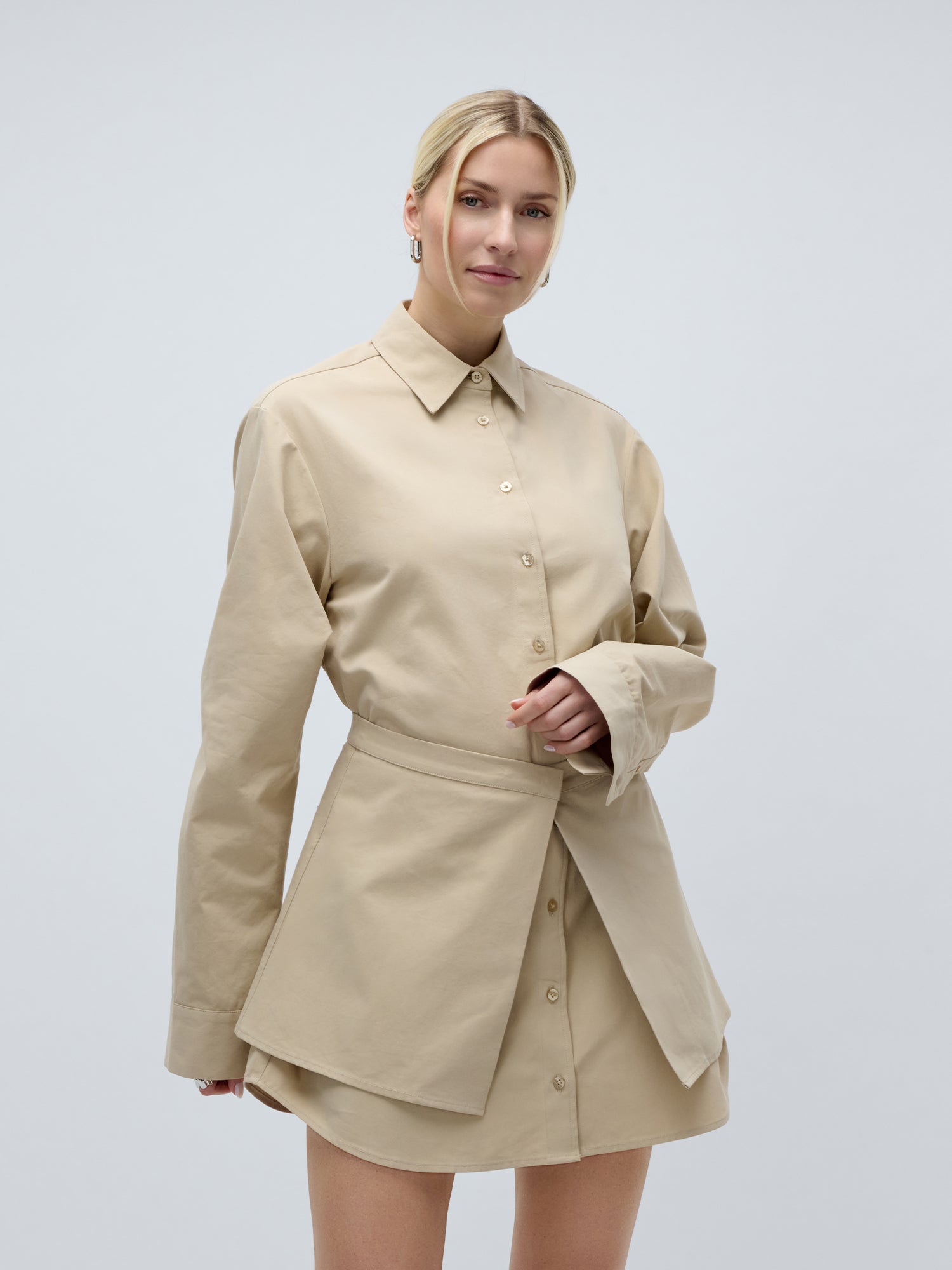 Eine Frau mit blonden Haaren, die zu einem tief sitzenden Dutt zusammengebunden sind, trägt das beigefarbene „Diana“-Kleid von LeGer by Lena Gercke mit langen Ärmeln, einem asymmetrischen, gestuften Rock, einem Kragen und einer lockeren Passform und steht vor einem schlichten hellgrauen Hintergrund.