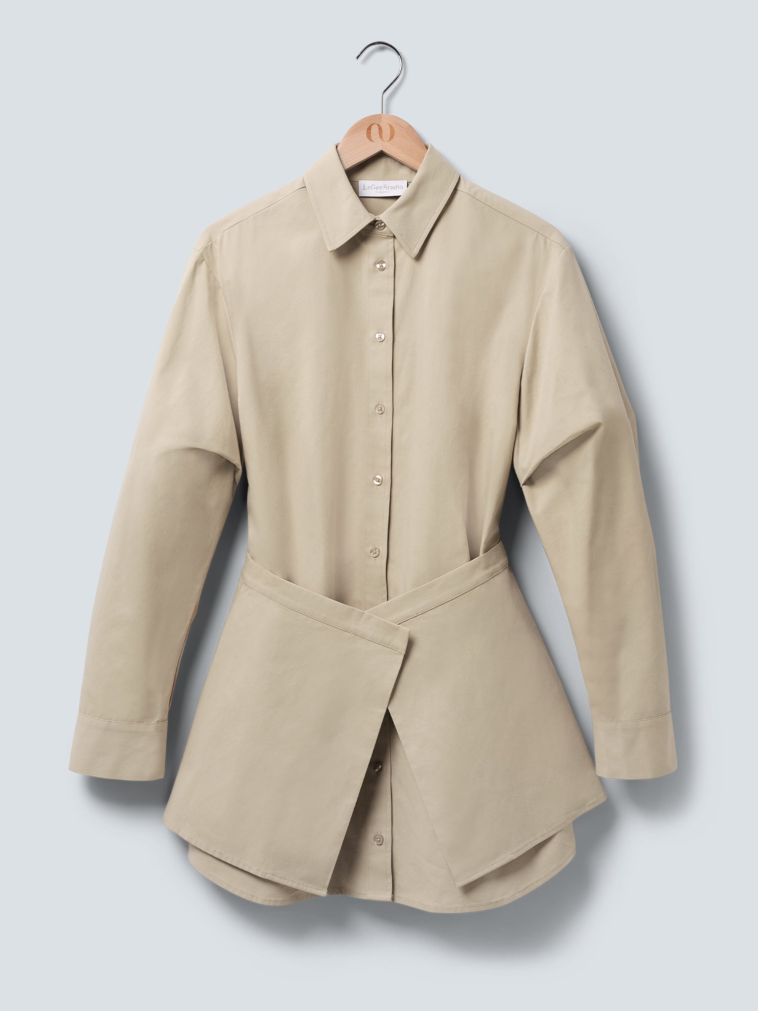 Das Kleid „Diana“ von LeGer by Lena Gercke ist ein beiges, langärmeliges Knopfkleid mit lockerer Passform und einem einzigartigen Wickeldetail an der Taille, das auf einem Holzkleiderbügel vor einem hellgrauen Hintergrund zu sehen ist.