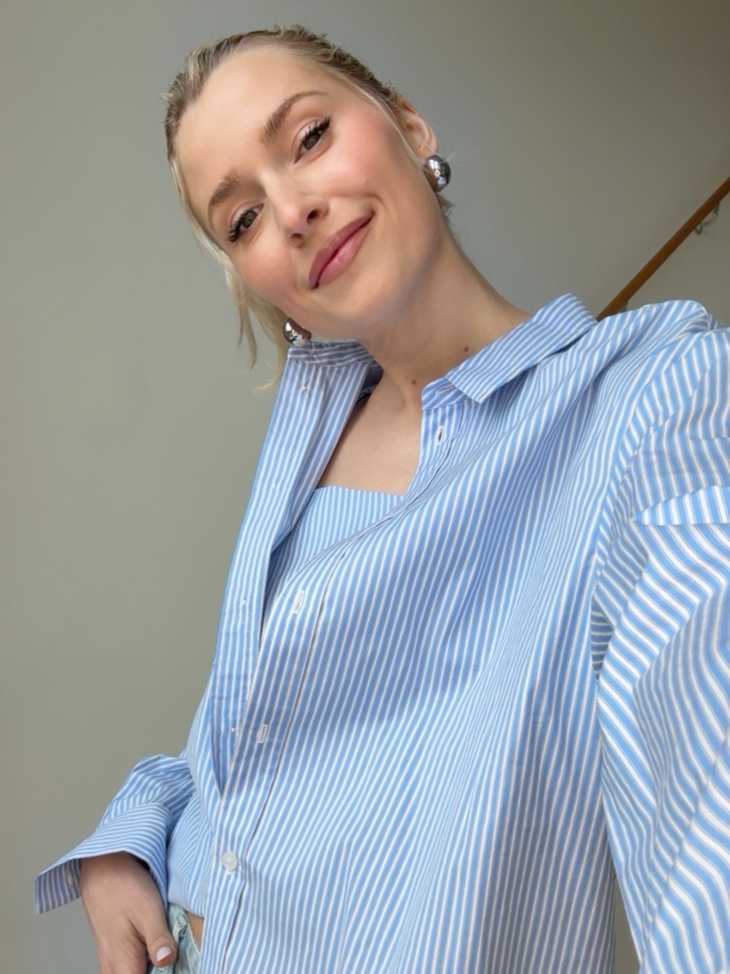 Eine lächelnde Frau mit blondem Haar trägt die Bluse Ginella von LeGer by Lena Gercke mit blau-weißem Streifenmuster und Hemdkragen, während sie für ein Selfie vor einem schlichten Hintergrund posiert.