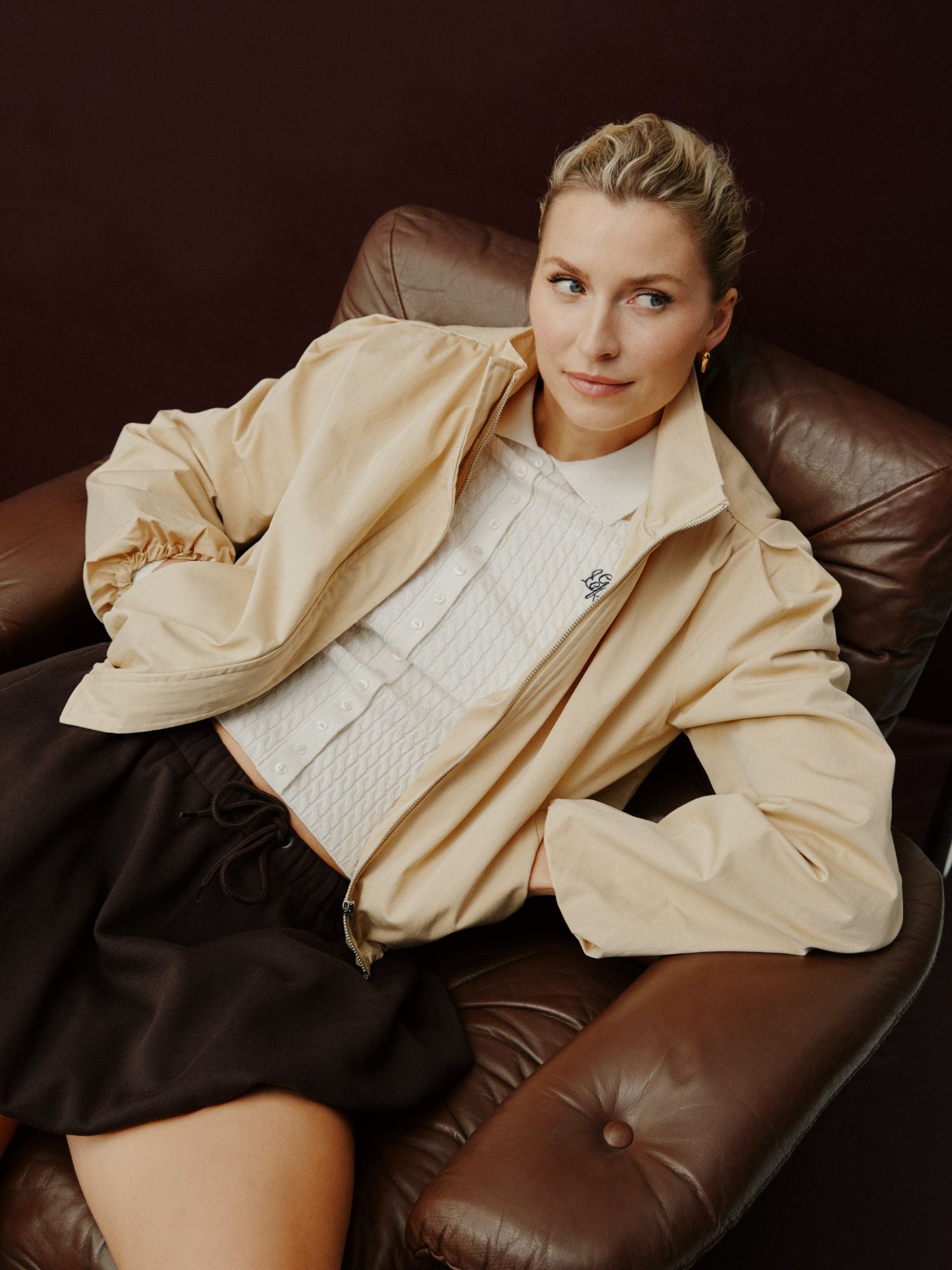 Eine Frau mit blonden Haaren sitzt auf einem braunen Ledersessel und trägt die hellbeige Romina Jacket von LeGer by Lena Gercke über einer weißen Bluse und einem dunklen Rock. Sie wirkt entspannt vor einem dunklen Hintergrund.