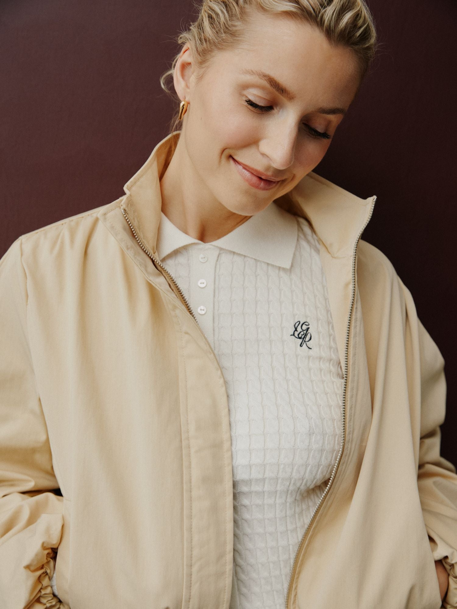 Eine Frau mit blonden Haaren, die Lena Gercke ähnelt, trägt eine hellbeige Jacke über einem weißen Manolja T-Shirt von LeGer by Lena Gercke, steht vor einem dunklen Hintergrund und schaut mit einem sanften Lächeln nach unten.