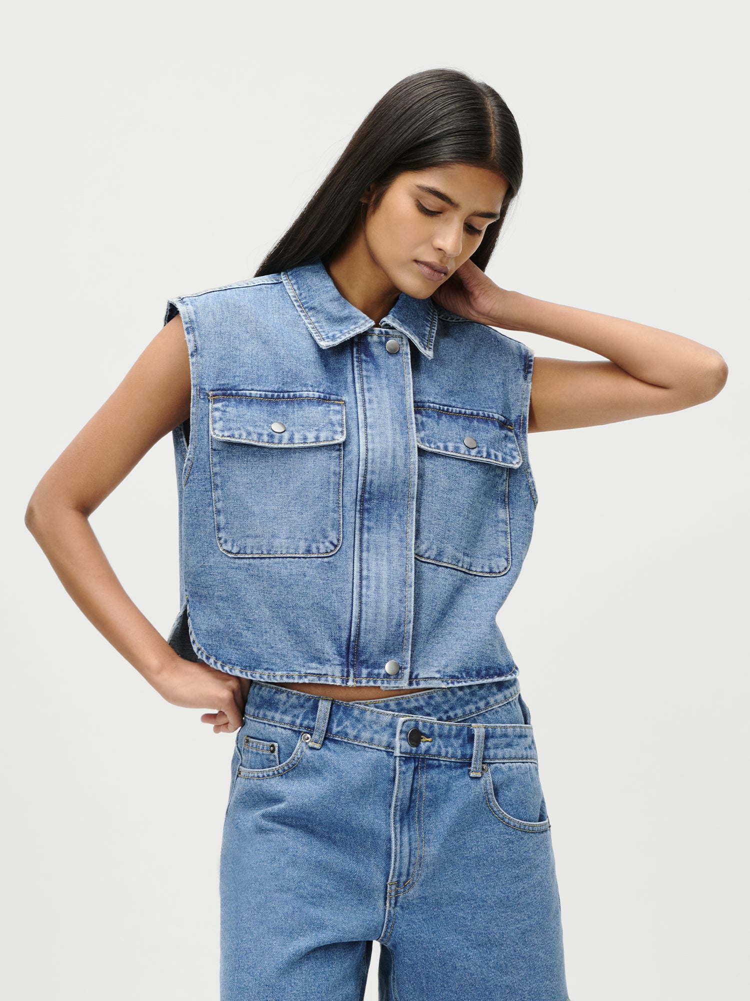 Eine Person mit langen, glatten dunklen Haaren trägt die Shirin Vest von LeGer by Lena Gercke in Größe 36. Die ärmellose Jeansweste hat große Vordertaschen und eine normale Passform. Sie posiert mit einer Hand im Nacken vor einem schlichten weißen Hintergrund.