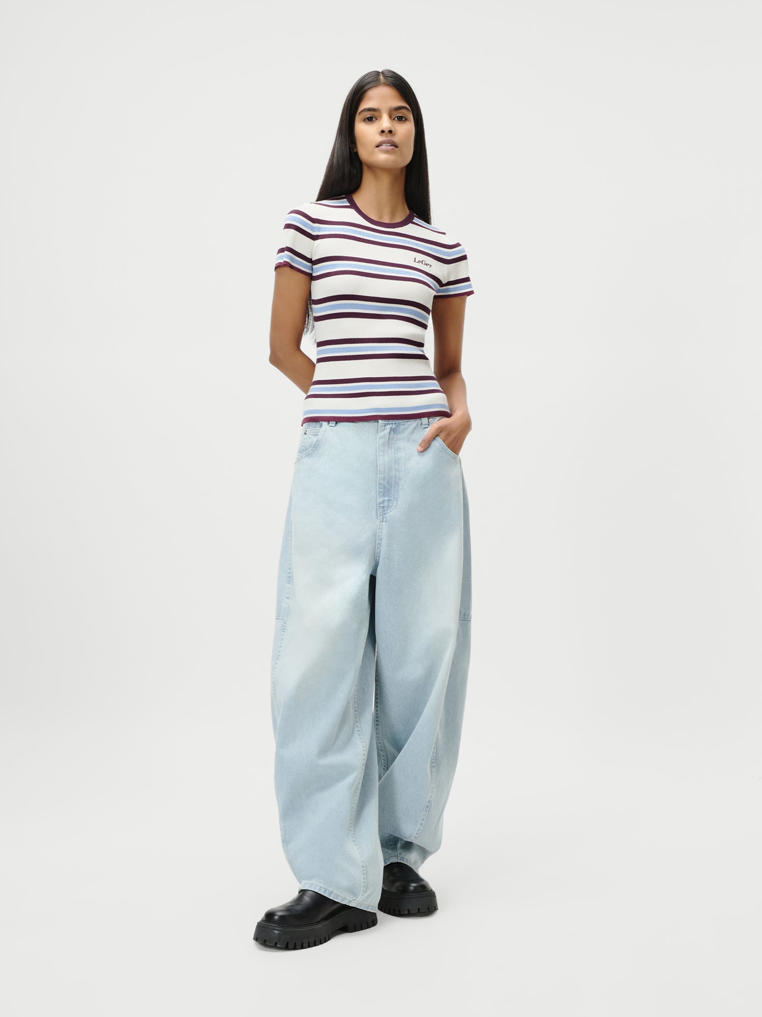 Eine Person steht mit den Händen in den Taschen vor einem weißen Hintergrund. Sie trägt ein tailliertes Danielle T-Shirt von LeGer by Lena Gercke im Streifendesign, gepaart mit einer hellblauen Jeans mit sehr weitem Bein und schwarzen, klobigen Schuhen.
