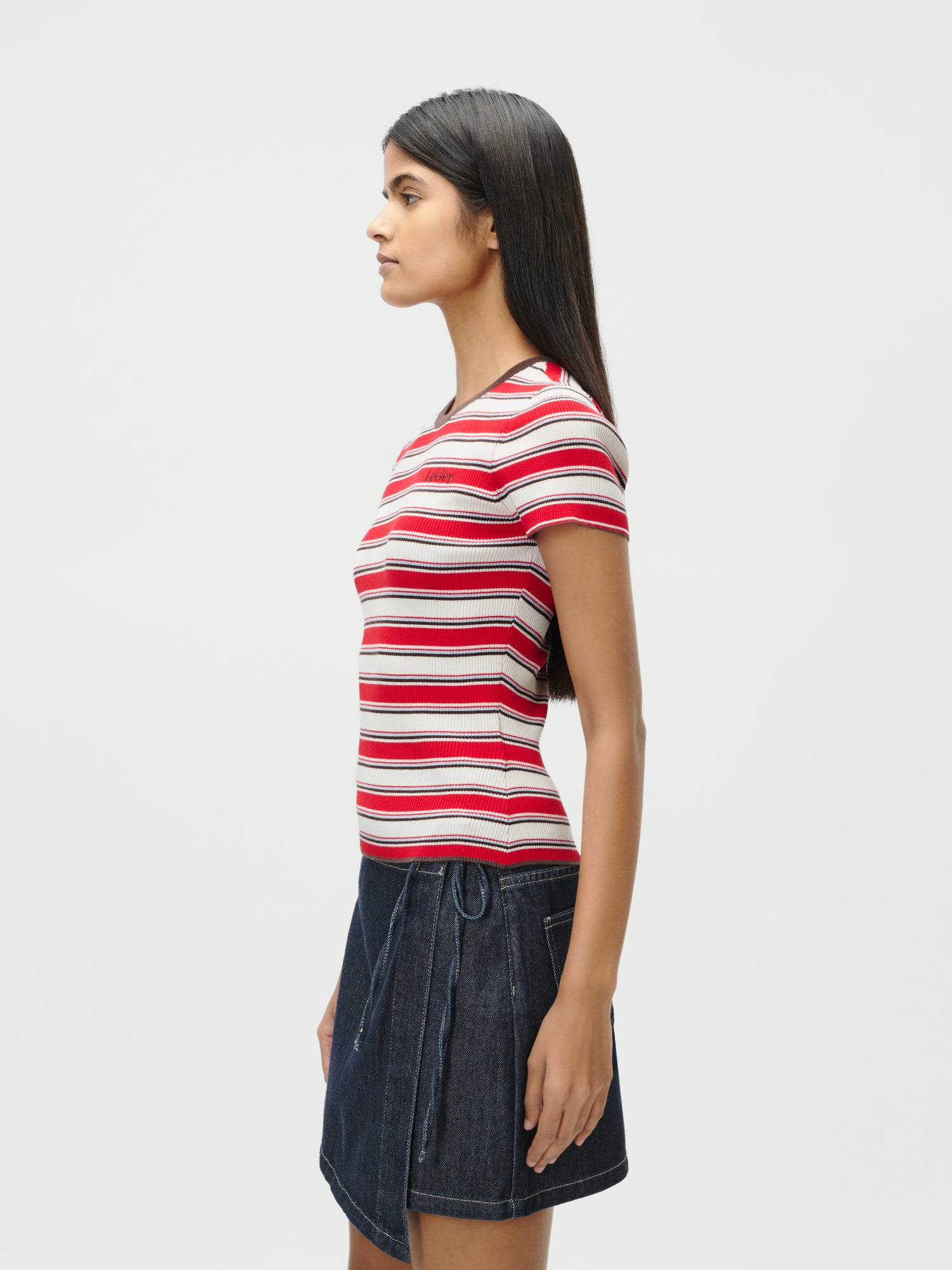 Eine Person mit langen dunklen Haaren steht im Profil vor einem schlichten weißen Hintergrund und trägt das rot-weiß-rosa gestreifte T-Shirt von LeGer by Lena Gercke Danielle sowie einen dunklen Jeansrock in Größe 36.