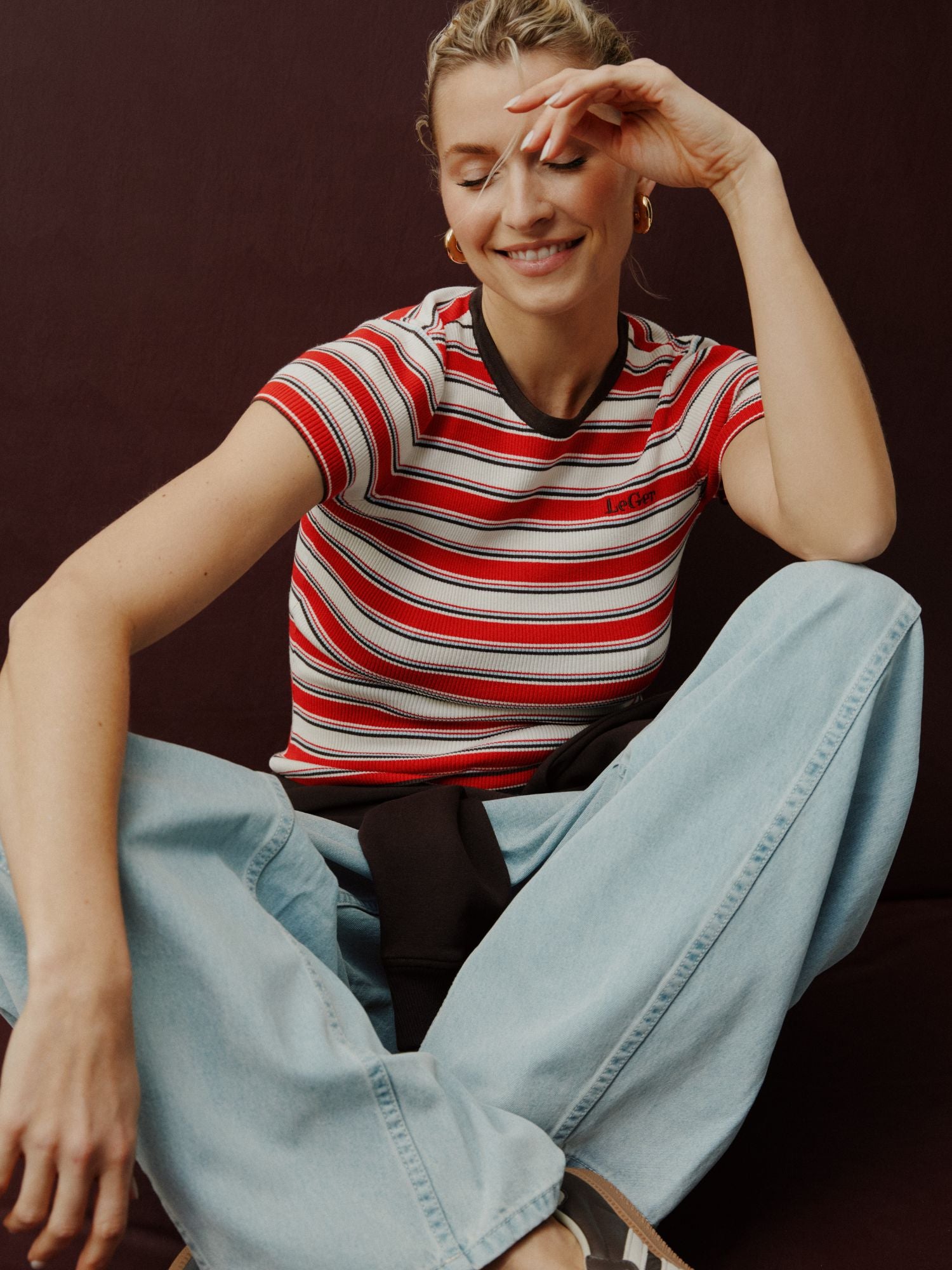 Eine Frau sitzt im Schneidersitz, lächelt mit geschlossenen Augen und einer Hand an der Stirn. Sie trägt das Danielle T-Shirt von LeGer by Lena Gercke in Größe 36 und hellblaue Jeans vor einem dunklen Hintergrund.