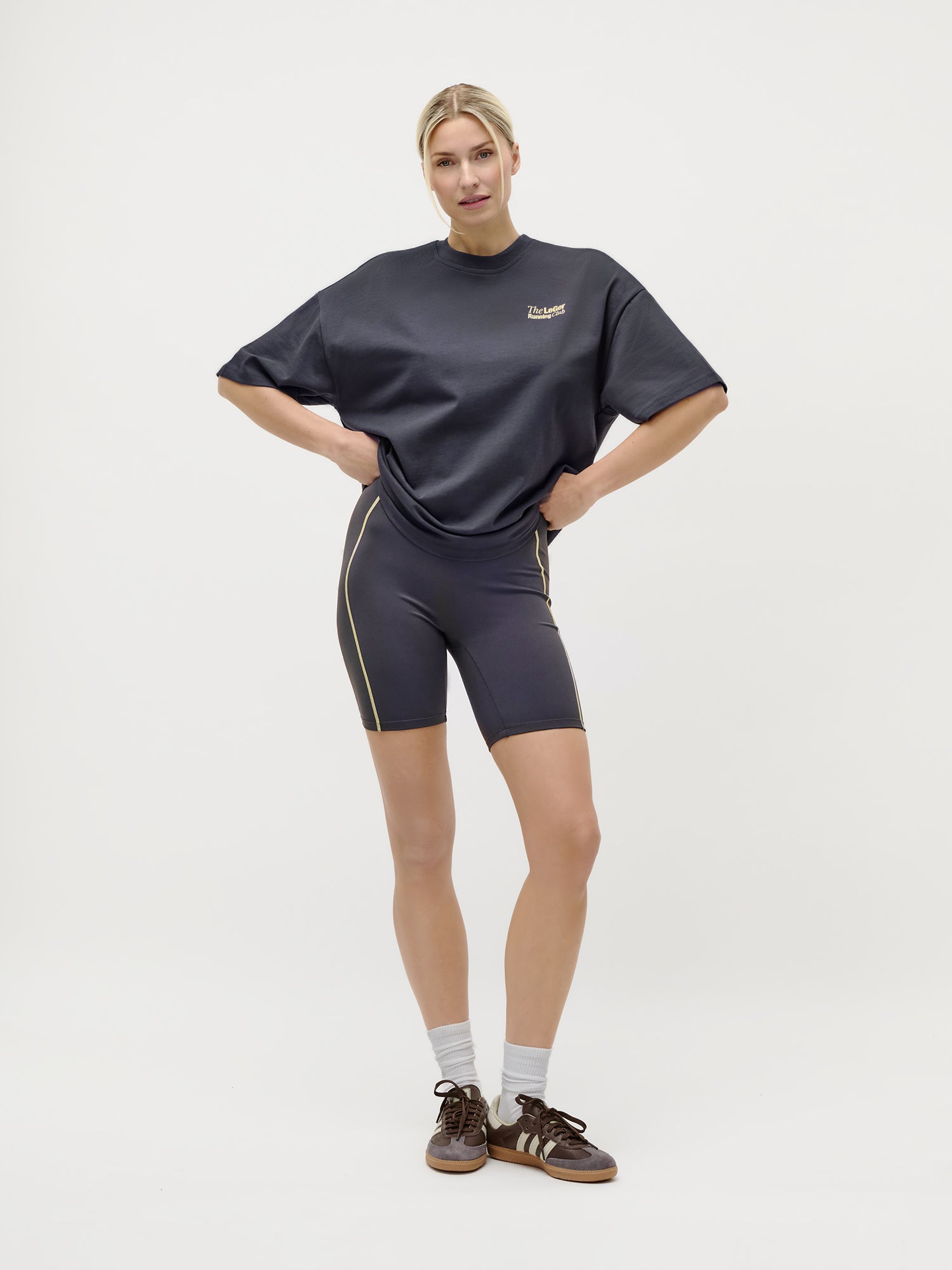 Eine Frau trägt ein übergroßes dunkelgraues T-Shirt „Regina“ von LeGer by Lena Gercke sowie Fahrradshorts mit goldenem Besatz, dazu weiße Socken und braune Sneaker – ein typischer Streetstyle-Look.