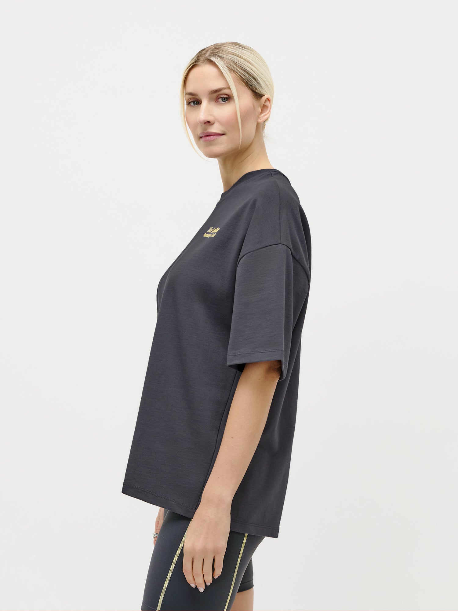 Eine Frau mit blonden Haaren, die zu einem tief sitzenden Dutt zusammengebunden sind, trägt das übergroße „Regina“-T-Shirt von LeGer by Lena Gercke sowie schwarze Shorts. Sie strahlt Streetstyle-Flair aus, während sie seitlich vor einem schlichten weißen Hintergrund steht und in die Kamera blickt.