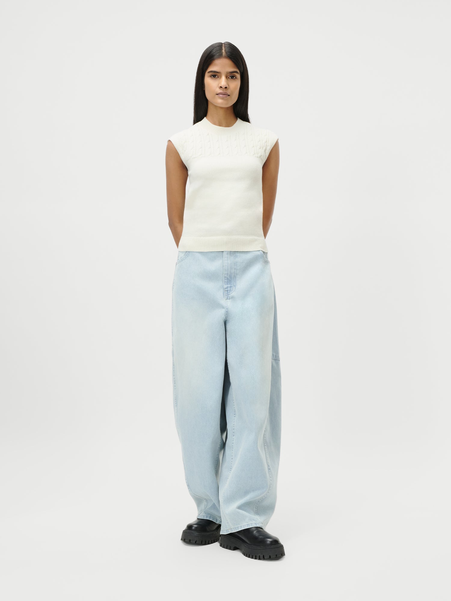 Eine Frau mit langen, glatten dunklen Haaren steht mit dem Gesicht nach vorne und den Armen hinter dem Rücken. Sie trägt eine Eilika-Hose von LeGer by Lena Gercke, gepaart mit schwarzen, klobigen Schuhen vor einem schlichten weißen Hintergrund.