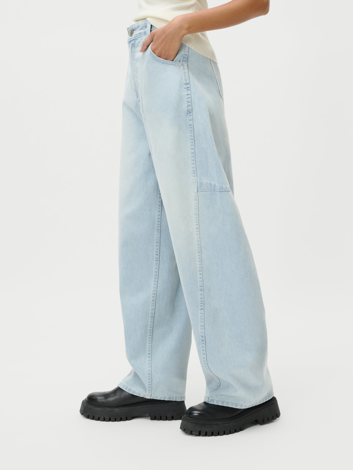 Diese Person trägt die Eilika Pants von LeGer by Lena Gercke. Sie kombiniert die Jeans mit hoher Taille und weitem Bein mit schwarzen, klobigen Plateauschuhen und einem leichten Top, die Hand in der Tasche vor einem schlichten weißen Hintergrund.