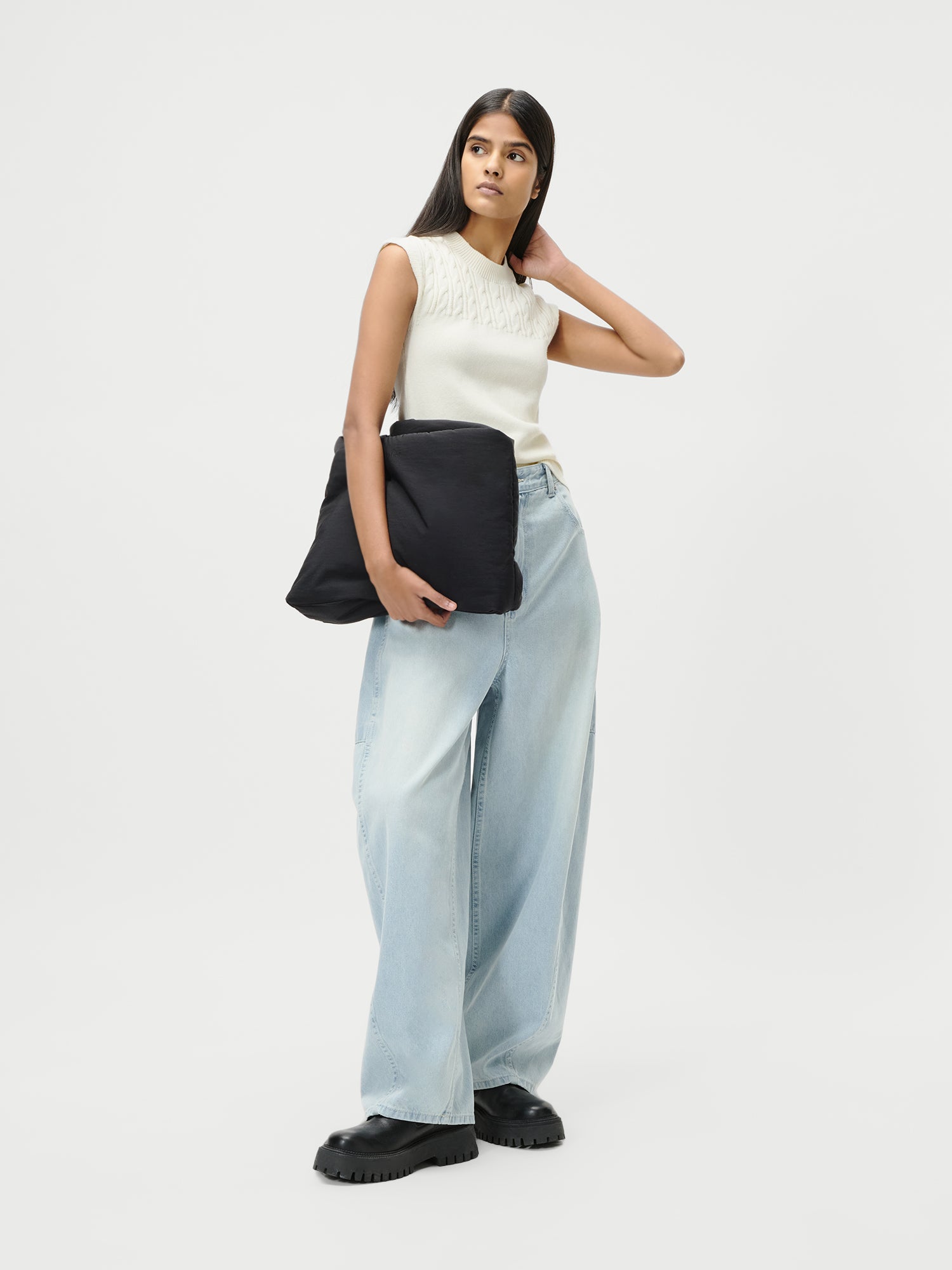 Eine Frau steht vor einem unifarbenen weißen Hintergrund und trägt die Eilika Pants von LeGer by Lena Gercke in Größe 36 - eine hellblaue Jeans mit weitem Bein - gepaart mit einem ärmellosen weißen Strickoberteil, schwarzen Schuhen und einer großen schwarzen kissenartigen Tasche.