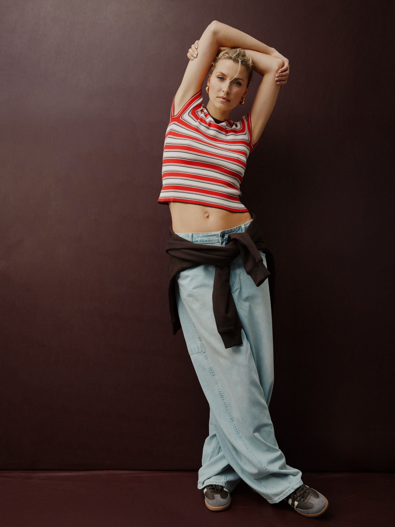 Das Model trägt die Eilika Pants von LeGer by Lena Gercke in Hellblau (Größe 36), kombiniert mit einem rot-weiß gestreiften Crop-Top, Turnschuhen und einem schwarzen Sweatshirt, das in der Taille gebunden wird, und posiert mit erhobenen Armen vor einem dunklen Hintergrund.