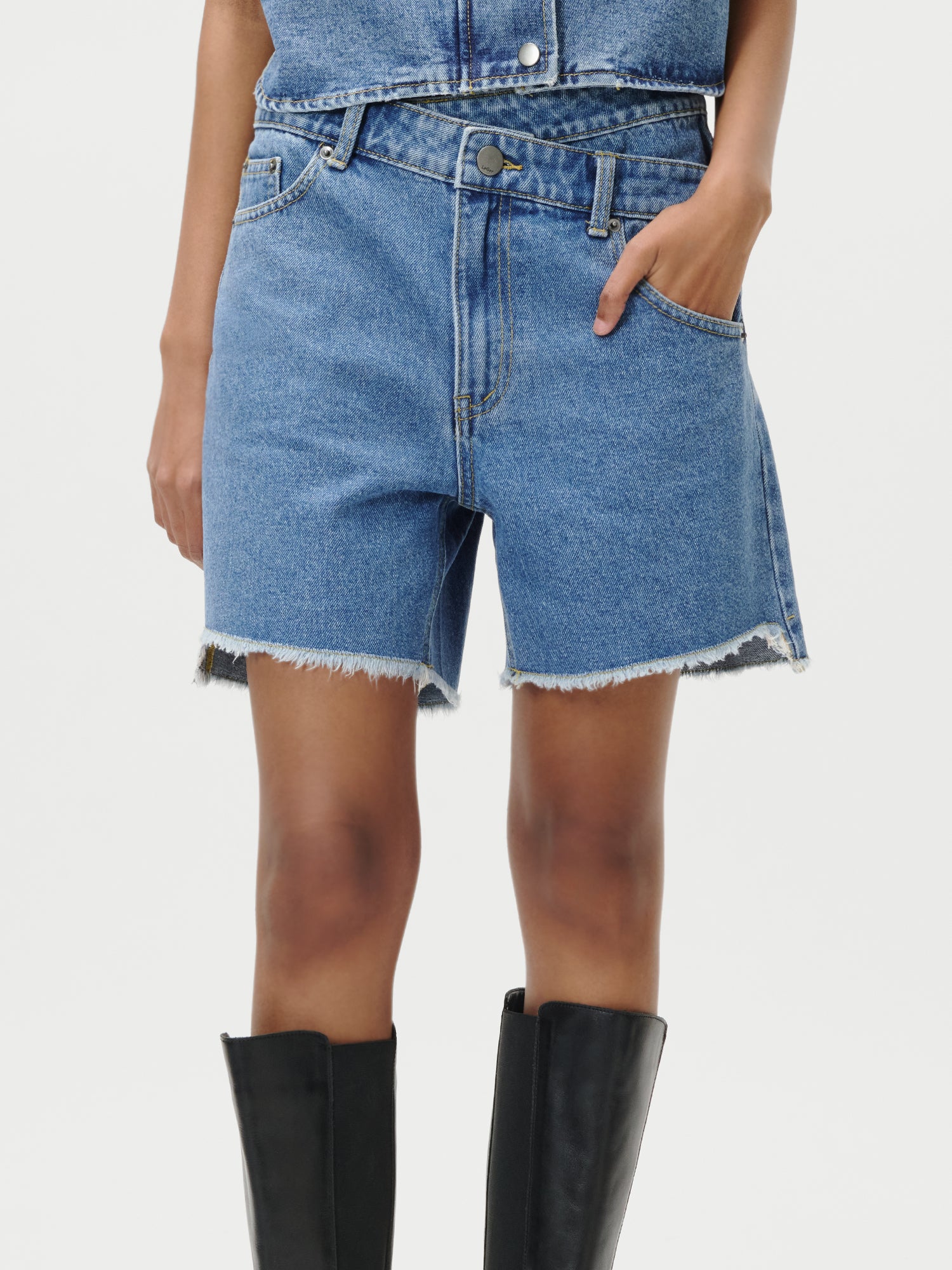 Ein Model trägt die Shirin Shorts von LeGer by Lena Gercke (Größe 36, halblang, ausgefranster Saum, blauer Denim) mit schwarzen kniehohen Stiefeln, eine Hand in der Tasche; das Foto zeigt sie von der Mitte des Oberkörpers bis zu den Knien vor einem weißen Hintergrund.
