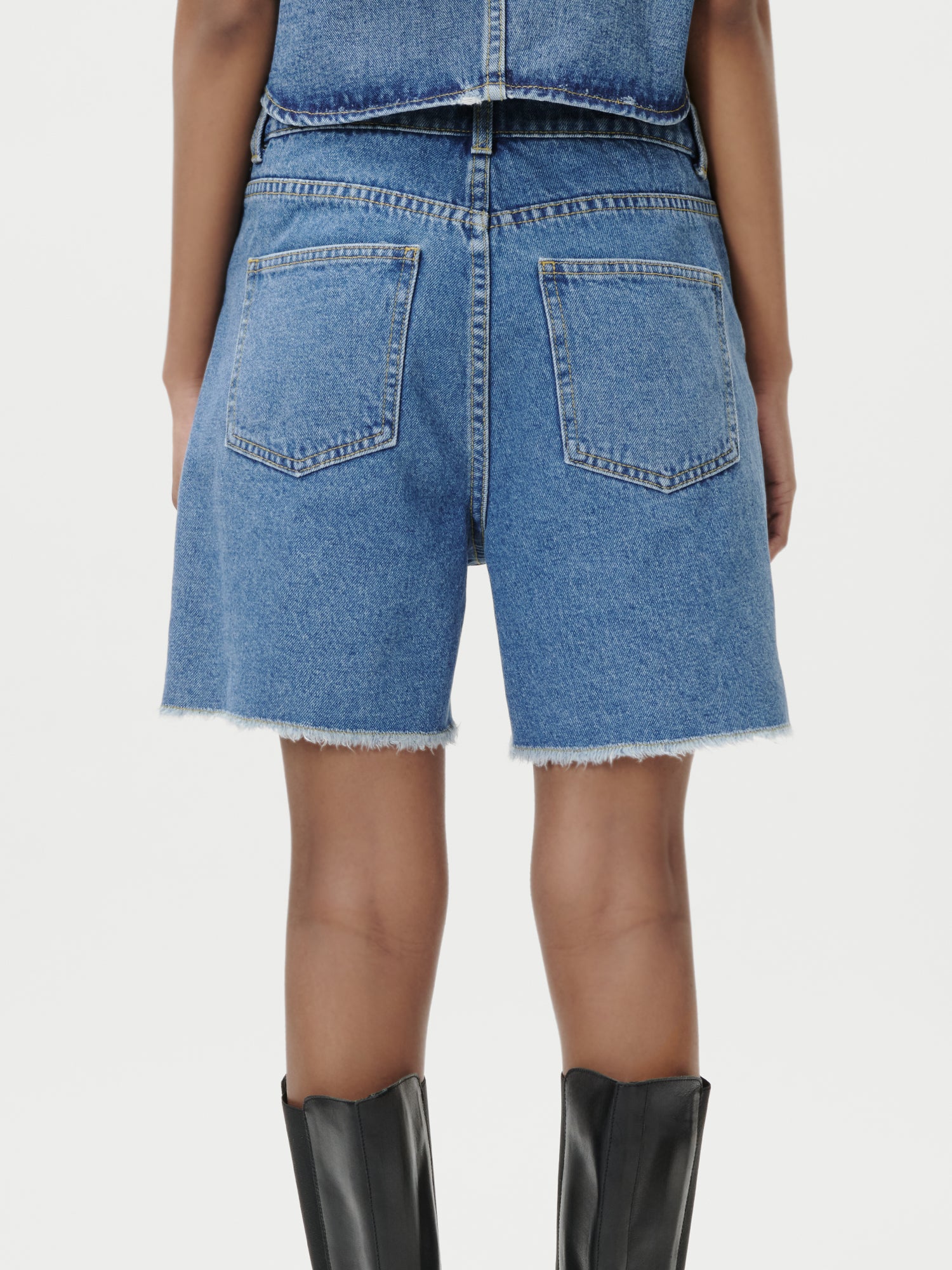 Model trägt LeGer by Lena Gercke Shirin Shorts aus blauem Denim mit ausgefransten Säumen und ein passendes Oberteil, von hinten gesehen. Kombiniert mit hohen schwarzen Stiefeln. Größe 36. Einfarbig weißer Hintergrund.