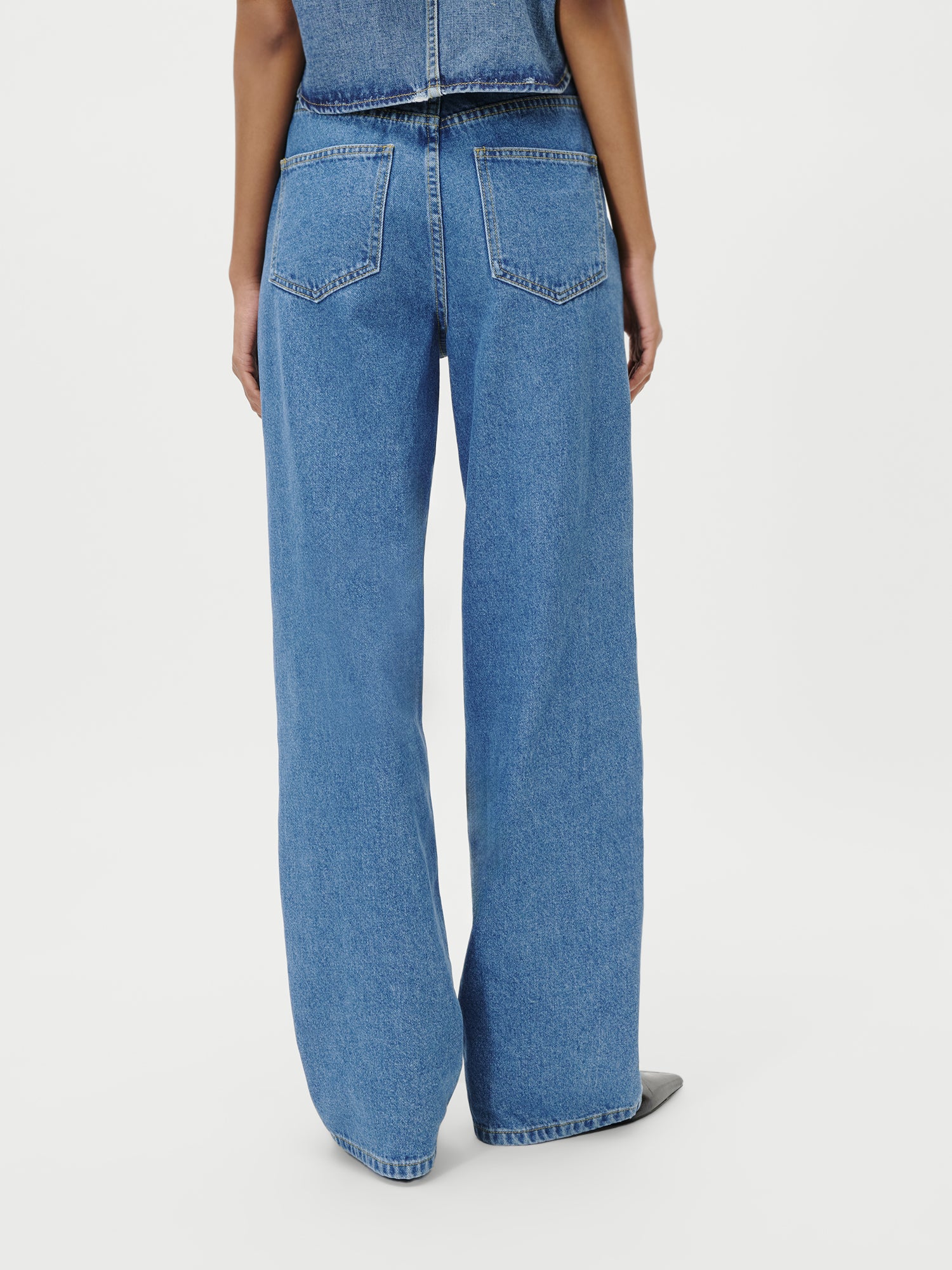Eine Person steht mit dem Gesicht zur Wand und trägt die Shirin Pants von LeGer by Lena Gercke - eine blaue Jeans mit hoher Taille und weitem Bein in Größe 36 mit sichtbaren Gesäßtaschen und schwarzen Schuhen - vor einem schlichten weißen Hintergrund.