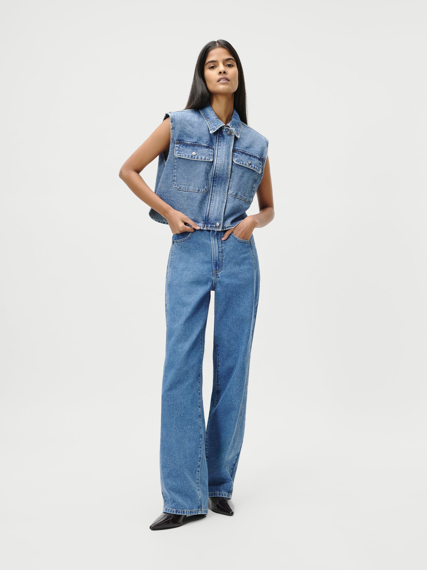 Eine Person steht selbstbewusst mit den Händen in den Hüften und trägt die Shirin Pants in Größe 36 von LeGer by Lena Gercke - eine blaue Jeans mit hoher Taille und weitem Bein - in Kombination mit einer ärmellosen Jeansweste vor einem schlichten weißen Hintergrund.
