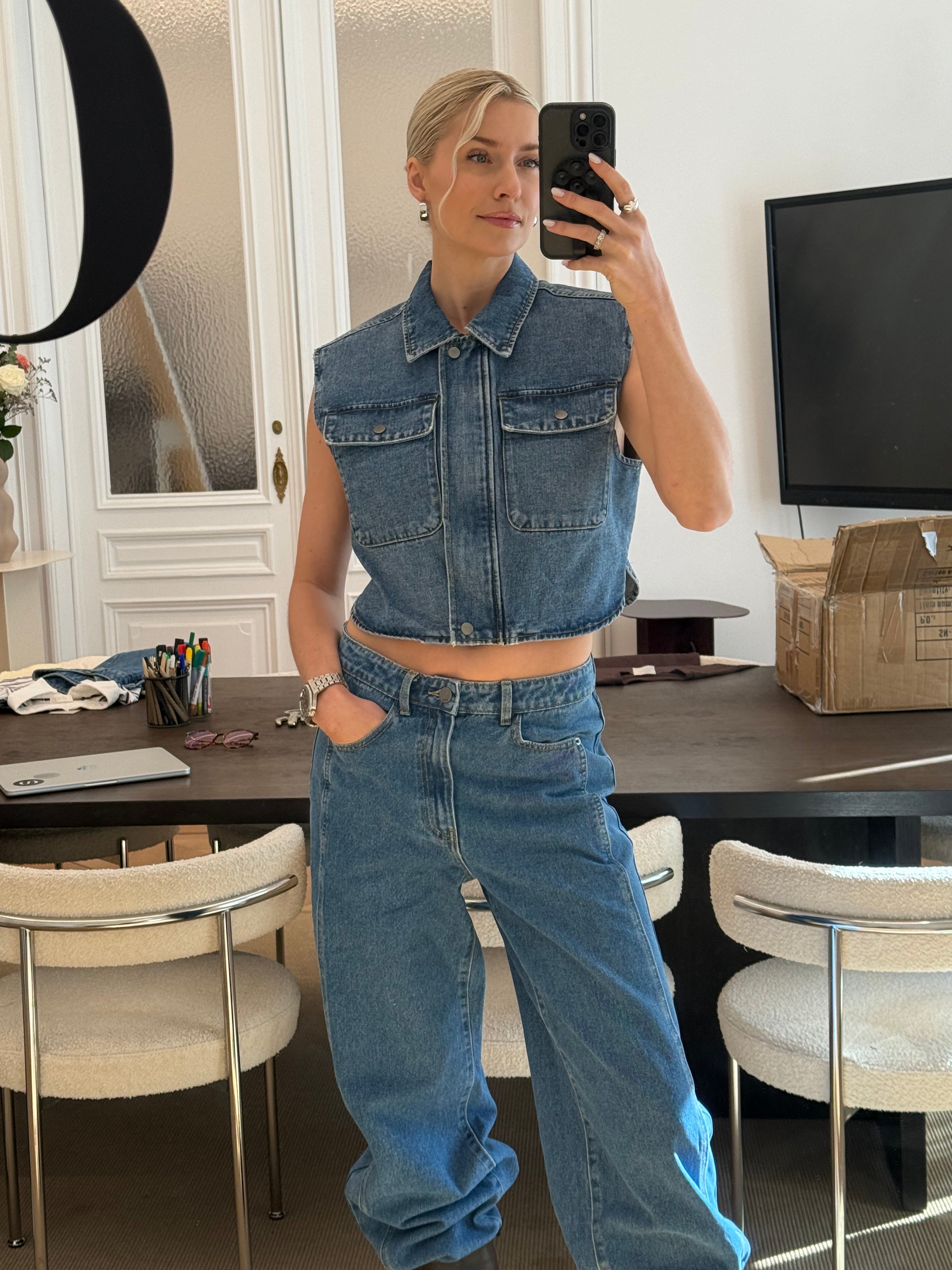 Eine Frau macht ein Selfie im Spiegel. Sie trägt eine Shirin Pants von LeGer by Lena Gercke und eine ärmellose Jeansweste, die schicke Damenmode unterstreicht. Hinter ihr stehen ein dunkler Tisch mit Stühlen, ein Fernseher und ein Pappkarton.