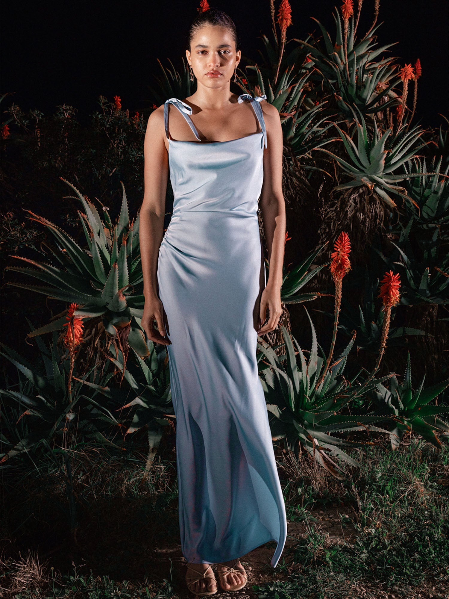 Eine Frau steht nachts im Freien vor großen stacheligen Sukkulenten mit roten Blüten. Sie trägt das Maxima Dress von LeGer by Lena Gercke - ein hellblaues, ärmelloses Maxikleid mit langem Schnitt und gebundenen Trägern - und Sandalen und schaut direkt in die Kamera.