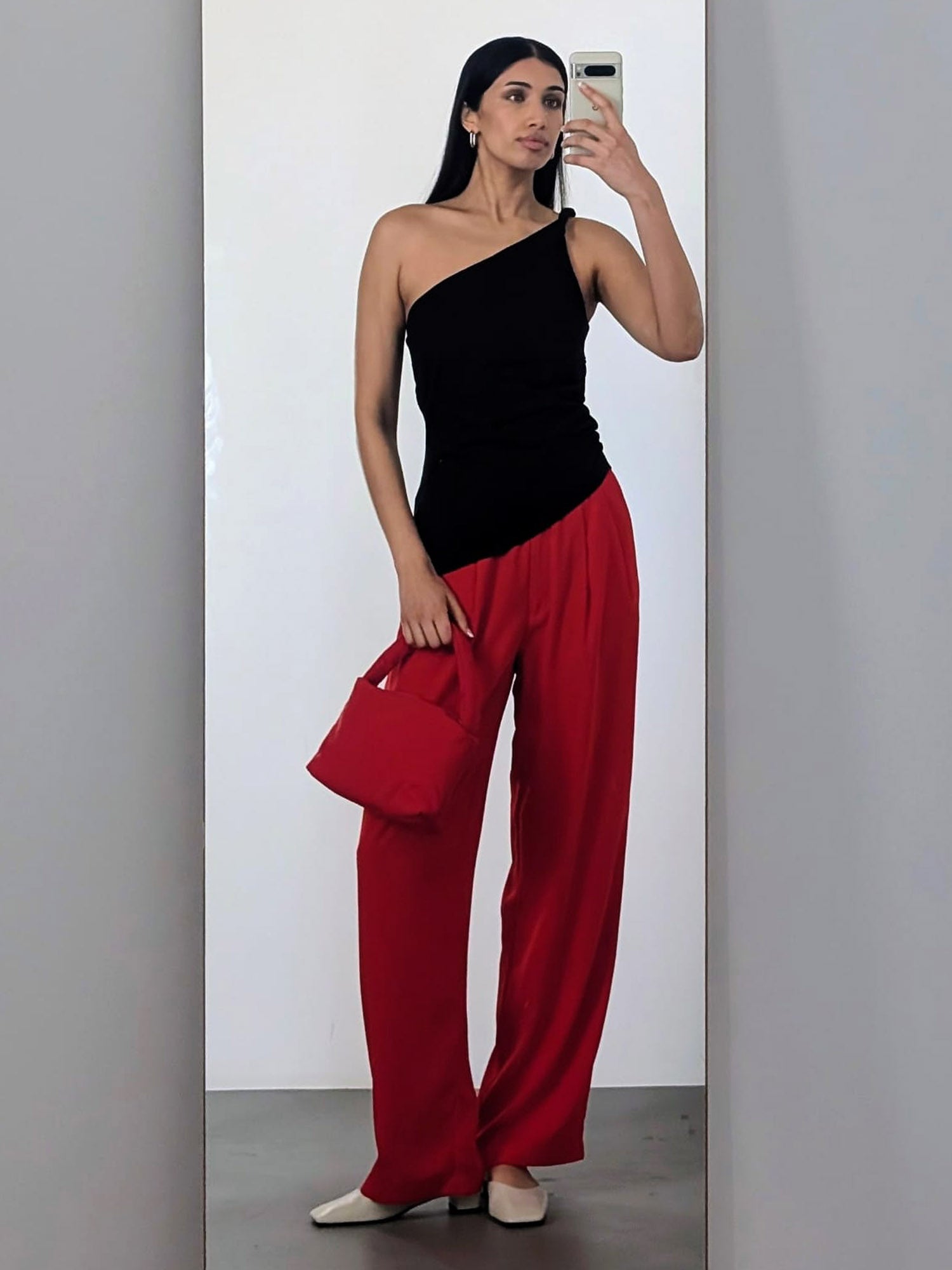 Eine Frau mit langen dunklen Haaren macht ein Spiegel-Selfie in dem LeGer by Lena Gercke Darleen Top (schmal, ärmellos, schwarz, One-Shoulder), einer weiten roten Hose (Größe 36), beigen Schuhen und einer roten Handtasche vor einem schlichten weißen Hintergrund.