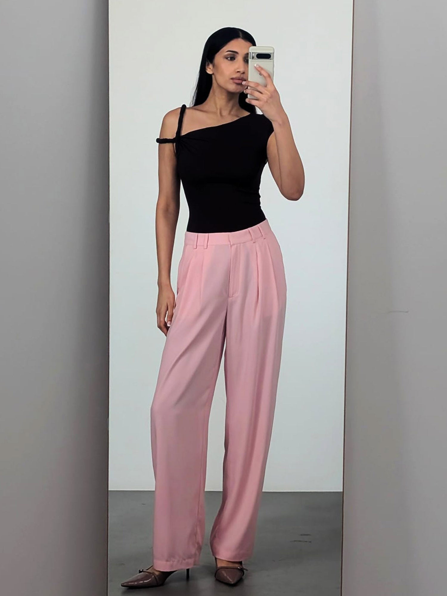 Eine Frau macht ein Selfie im Spiegel und trägt das Fenna Top von LeGer by Lena Gercke, ein schwarzes, schmal geschnittenes, ärmelloses One-Shoulder-Design, kombiniert mit einer hoch taillierten, lockeren rosa Hose in Größe 36 und spitzen Absätzen. Der Hintergrund ist minimalistisch und neutral.