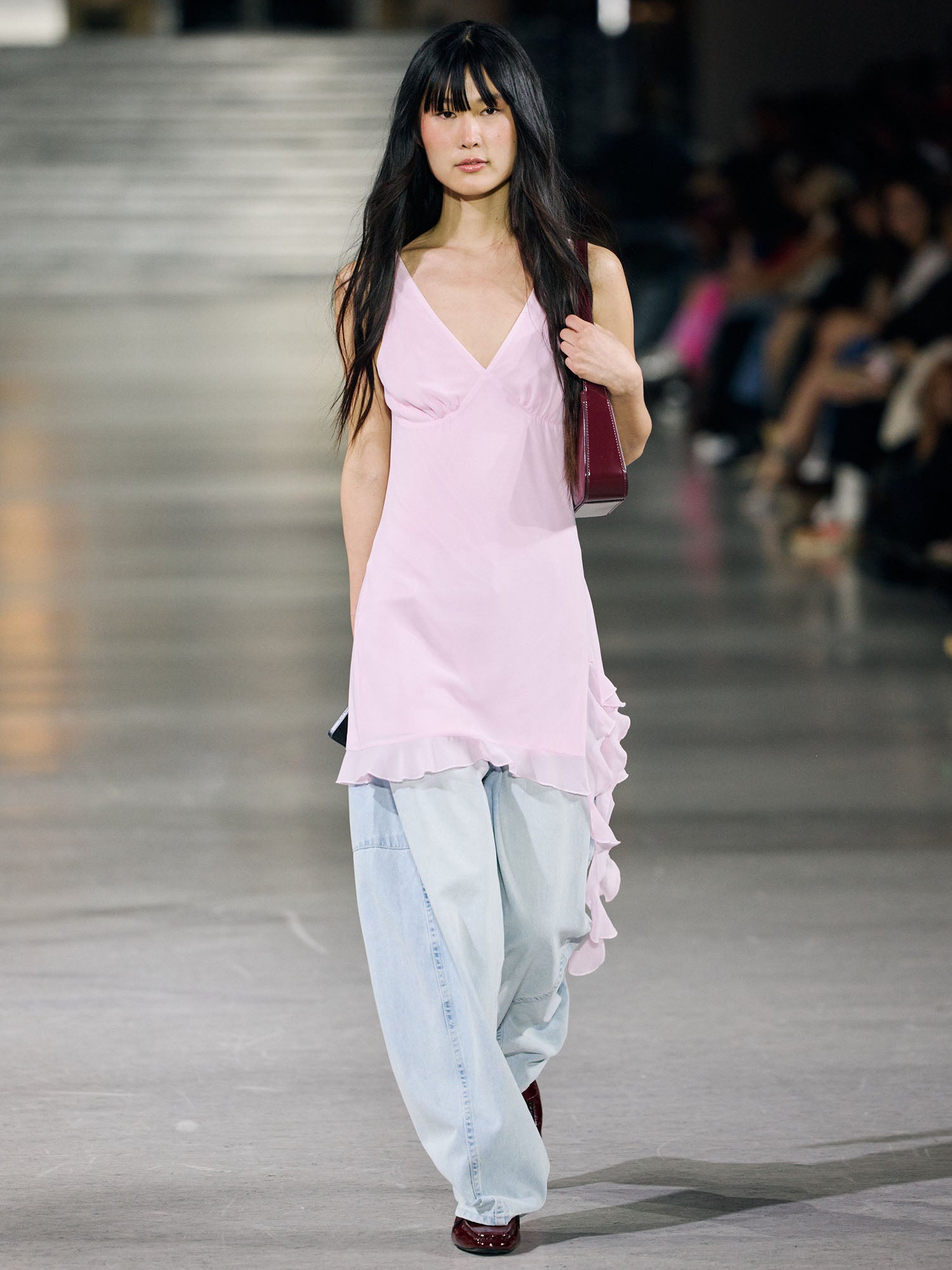 Ein Model läuft über den Laufsteg in dem Hilka-Kleid von LeGer by Lena Gercke, einem ärmellosen hellrosa Minikleid, das über eine weite hellblaue Jeans gestylt ist. Sie hat langes dunkles Haar mit Pony und trägt eine weinrote Tasche, während im Hintergrund Schaulustige sitzen.
