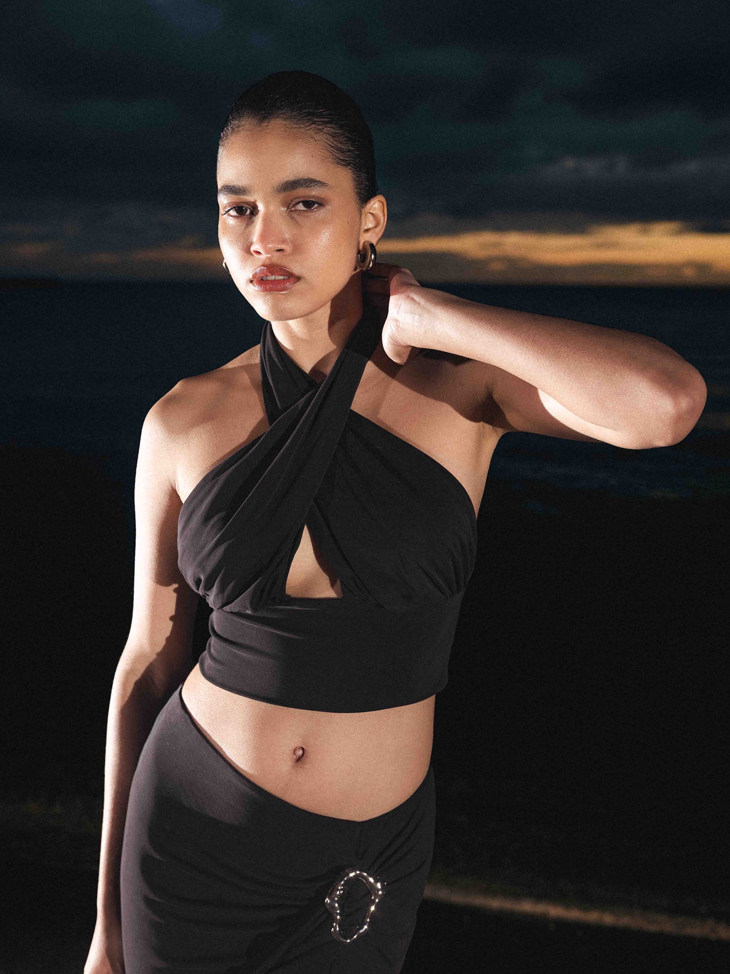 Bei Sonnenuntergang posiert eine Frau im Freien und trägt dabei das LeGer by Lena Gercke Agathe Top, ein figurbetontes, ärmelloses Crossover-Neckholder-Top, das mit einem passenden Rock und einer dekorativen Schnalle kombiniert ist. Sie posiert mit hochgestecktem Haar vor einem dunklen Hintergrund mit dezenter Beleuchtung.
