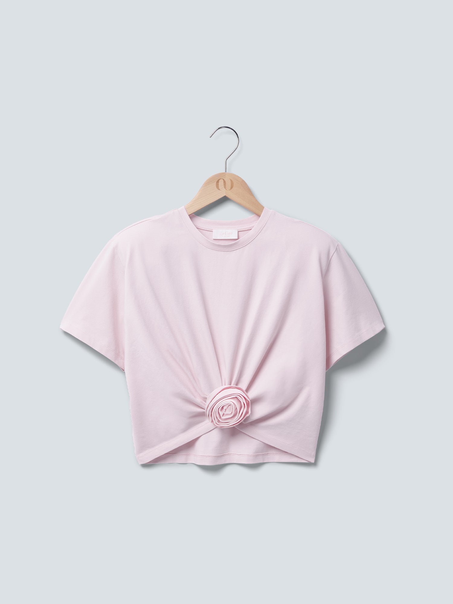 Das „Ina“-T-Shirt von LeGer by Lena Gercke ist ein hellrosa, kurzärmeliges Oberteil mit Rundhalsausschnitt, geknoteter Vorderseite und Rosendetail, das auf einem Holzkleiderbügel vor grauem Hintergrund abgebildet ist.