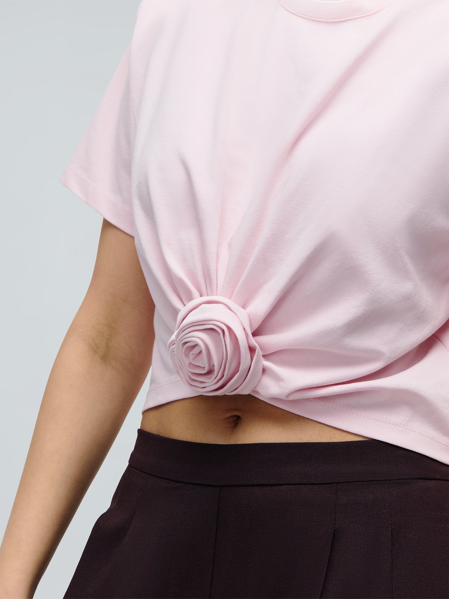 Eine Person trägt das „Ina“-T-Shirt von LeGer by Lena Gercke – dessen geknotete Vorderseite eine Rosenform bildet – kombiniert mit einer schwarzen Hose mit hoher Taille. Das Foto zeigt den Oberkörper und einen Teil eines Arms.