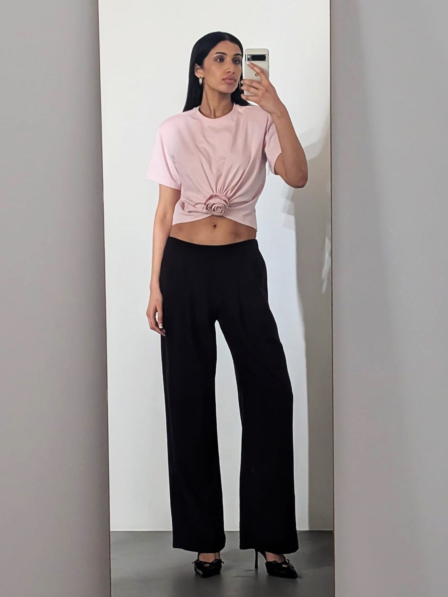 Eine Frau macht ein Spiegel-Selfie. Sie trägt das T-Shirt „Ina“ von LeGer by Lena Gercke mit Rundhalsausschnitt, das an der Taille geknotet ist, dazu eine schwarze Hose mit weitem Bein und schwarze Pumps. Der Hintergrund ist schlicht und neutral.