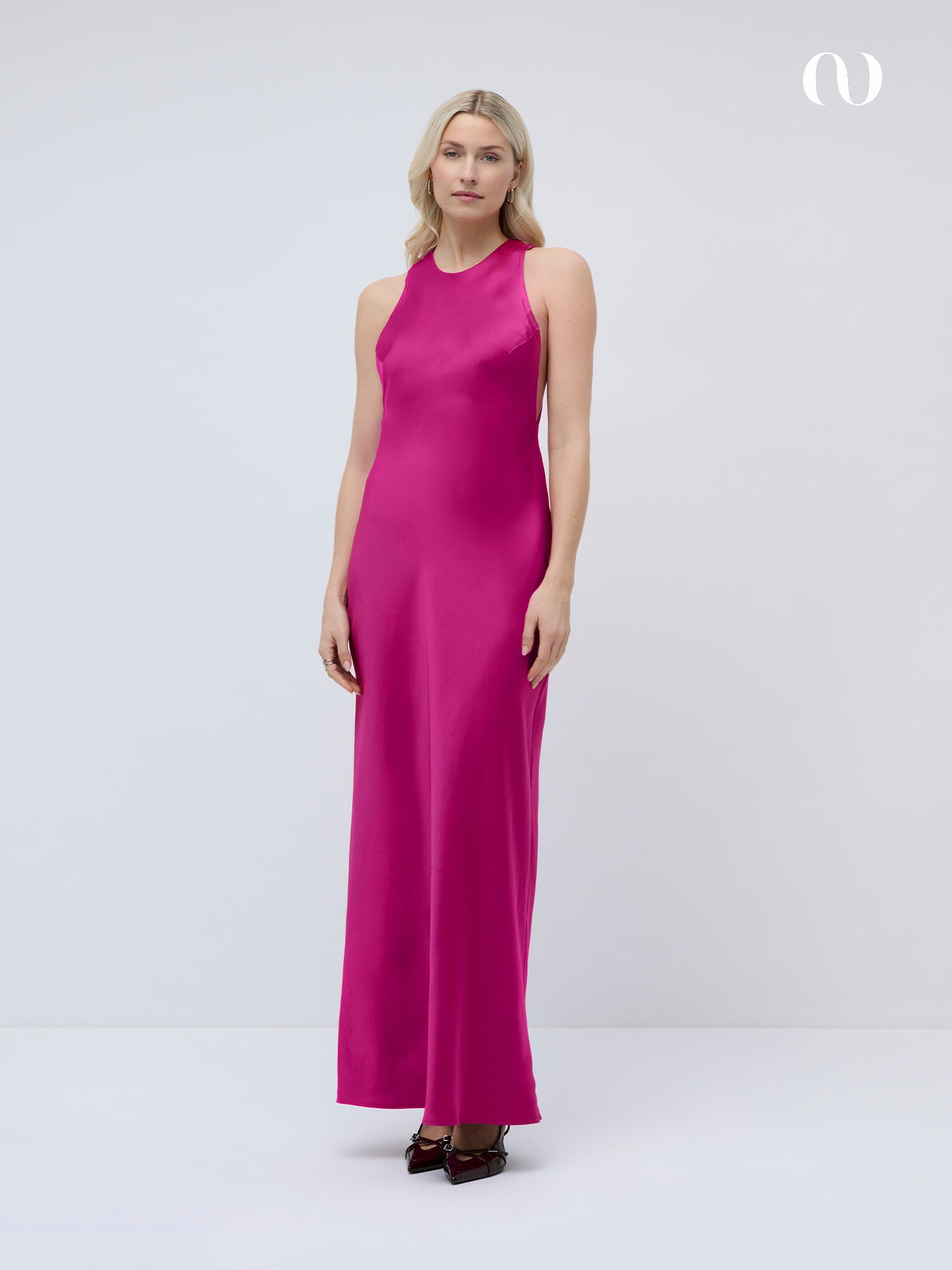 Eine Frau mit langen blonden Haaren trägt das Else Dress by LeGer by Lena Gercke, ein ärmelloses, bodenlanges Maxikleid aus leuchtend rosa Satin, gepaart mit dunklen Absätzen, vor einem schlichten weißen Hintergrund.