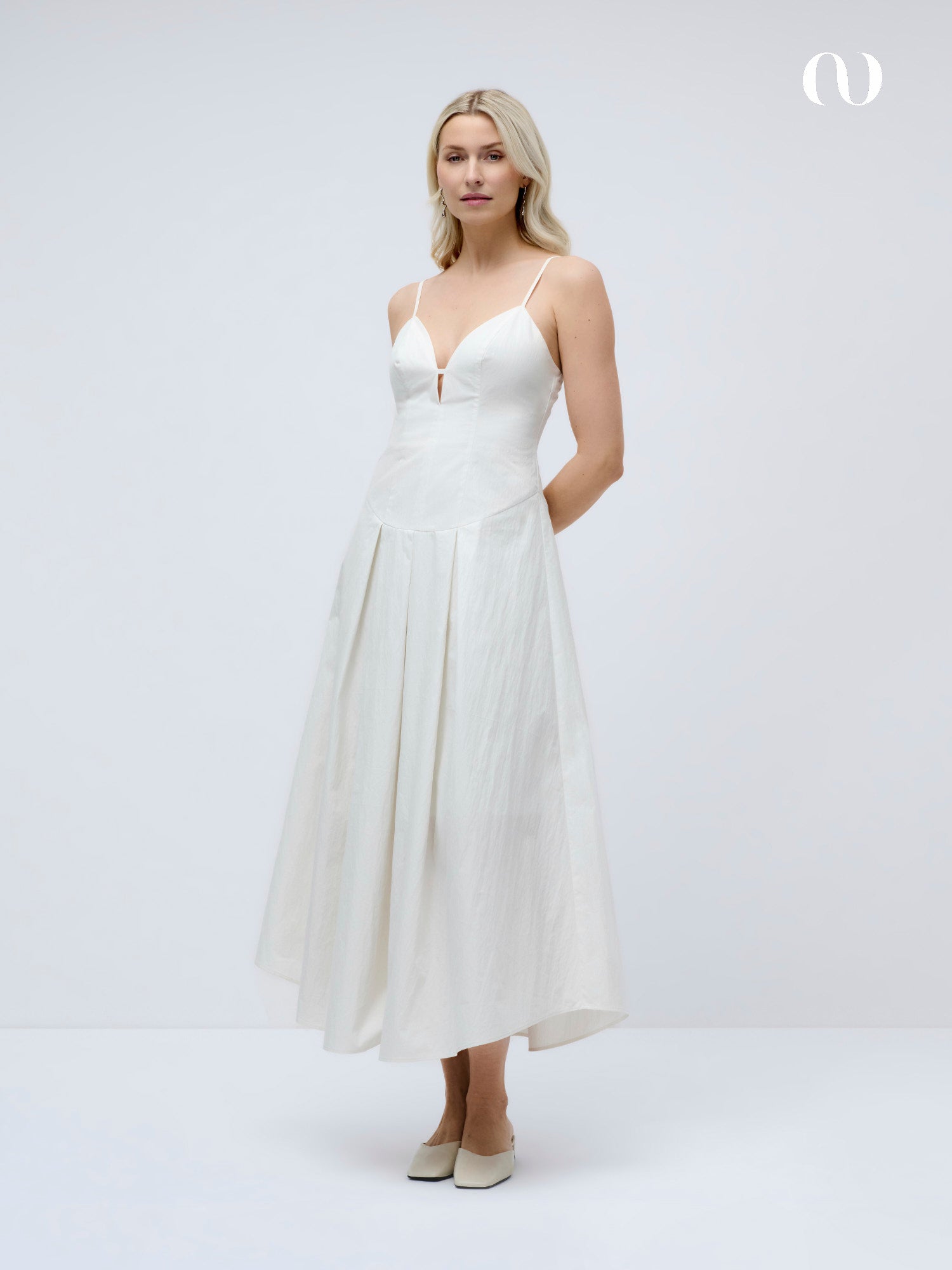 Eine Frau mit blondem Haar trägt das LeGer by Lena Gercke Jella Dress, ein weißes, ärmelloses, knöchellanges Kleid mit dünnen Trägern, V-Ausschnitt und Kellerfalten-Akzenten. Sie steht mit hellen Schuhen vor einem schlichten weißen Hintergrund.