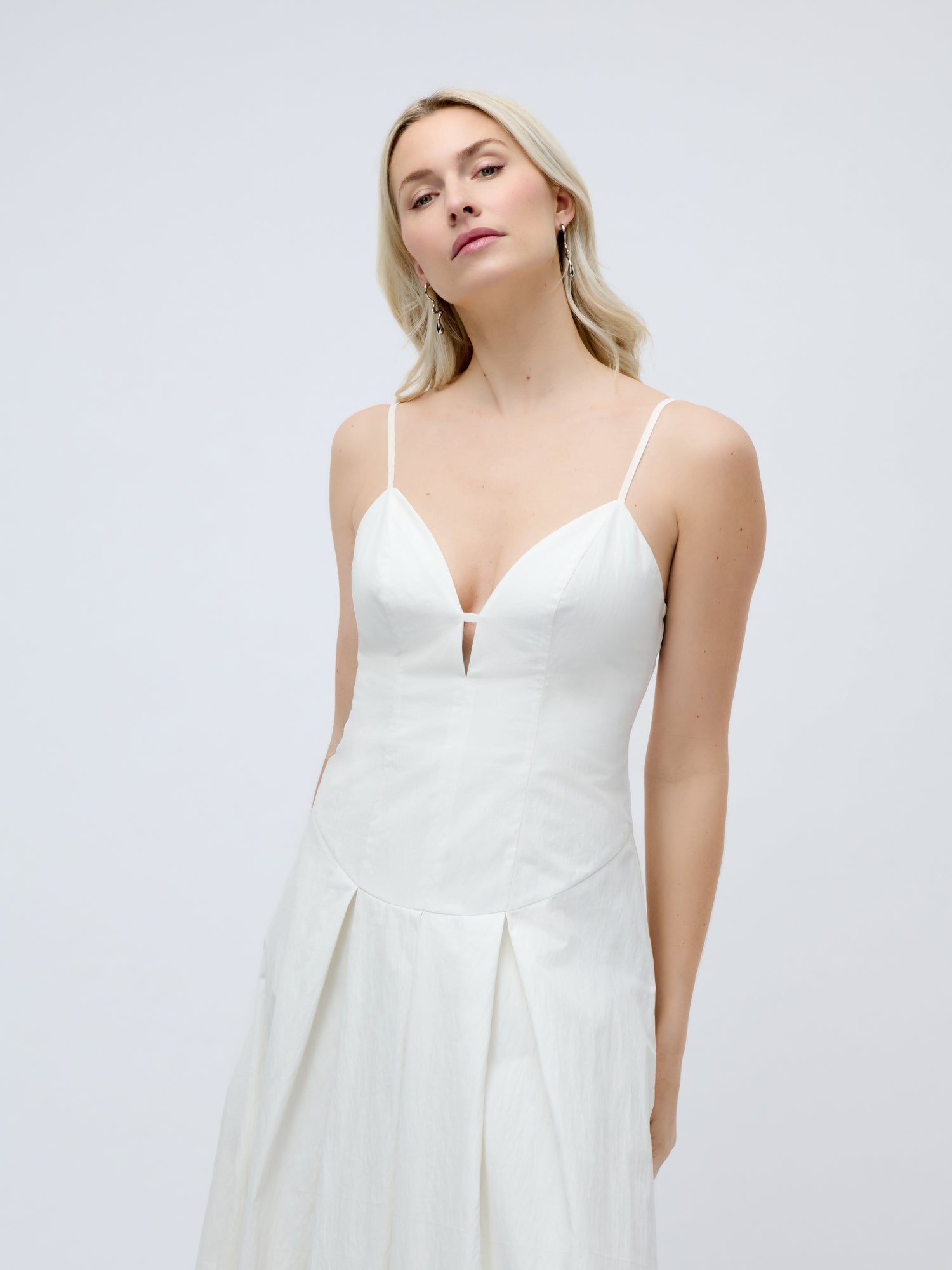 Eine Frau mit langen blonden Haaren präsentiert das Kleid „Jella“ von LeGer by Lena Gercke – ein ärmelloses, weißes, knöchellanges Kleid mit schmalen Trägern und einem tiefen V-Ausschnitt –, während sie vor einem schlichten, hellen Hintergrund steht.