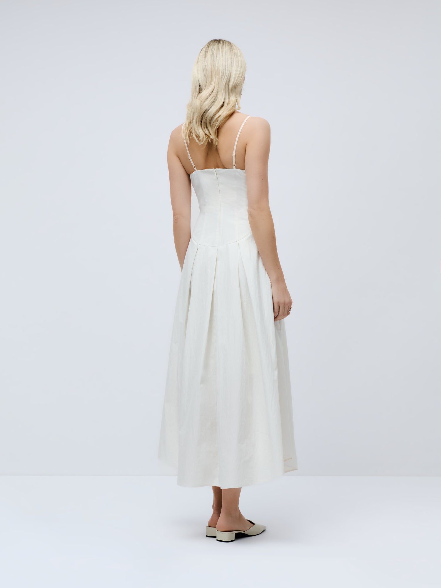 Eine Frau mit blonden Haaren, die mit dem Rücken zur Kamera steht, trägt das knöchellange „Jella“-Kleid von LeGer by Lena Gercke – ein weißes, elegantes Kleid mit dünnen Trägern – in einem gut beleuchteten Studio vor weißem Hintergrund und dazu flache beige Schuhe.
