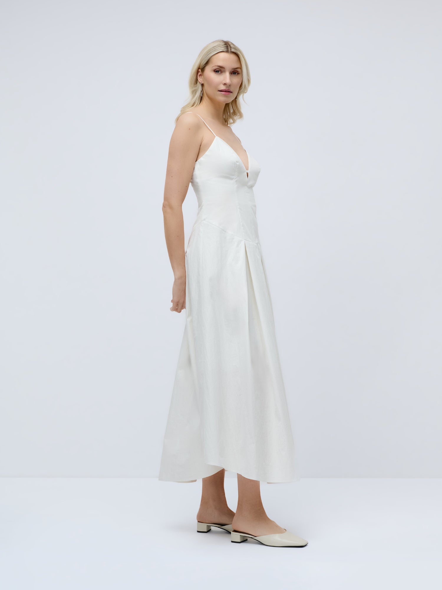 Eine blonde Frau präsentiert das Kleid „Jella“ von LeGer by Lena Gercke – ein weißes, knöchellanges, elegantes Kleid mit schmalen Trägern und V-Ausschnitt –, kombiniert mit cremefarbenen Pantoletten mit niedrigem Absatz vor einem hellgrauen Hintergrund.