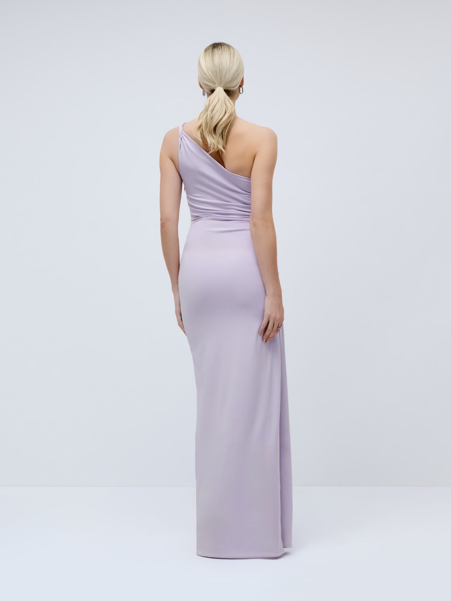 Eine Frau mit blonden Haaren, die zu einem tief sitzenden Pferdeschwanz gebunden sind, präsentiert das elegante „Claire“-Kleid von LeGer by Lena Gercke – ein hellviolettes Ein-Schulter-Modell – und steht mit dem Rücken zur Kamera vor einem schlichten, hellen Hintergrund.