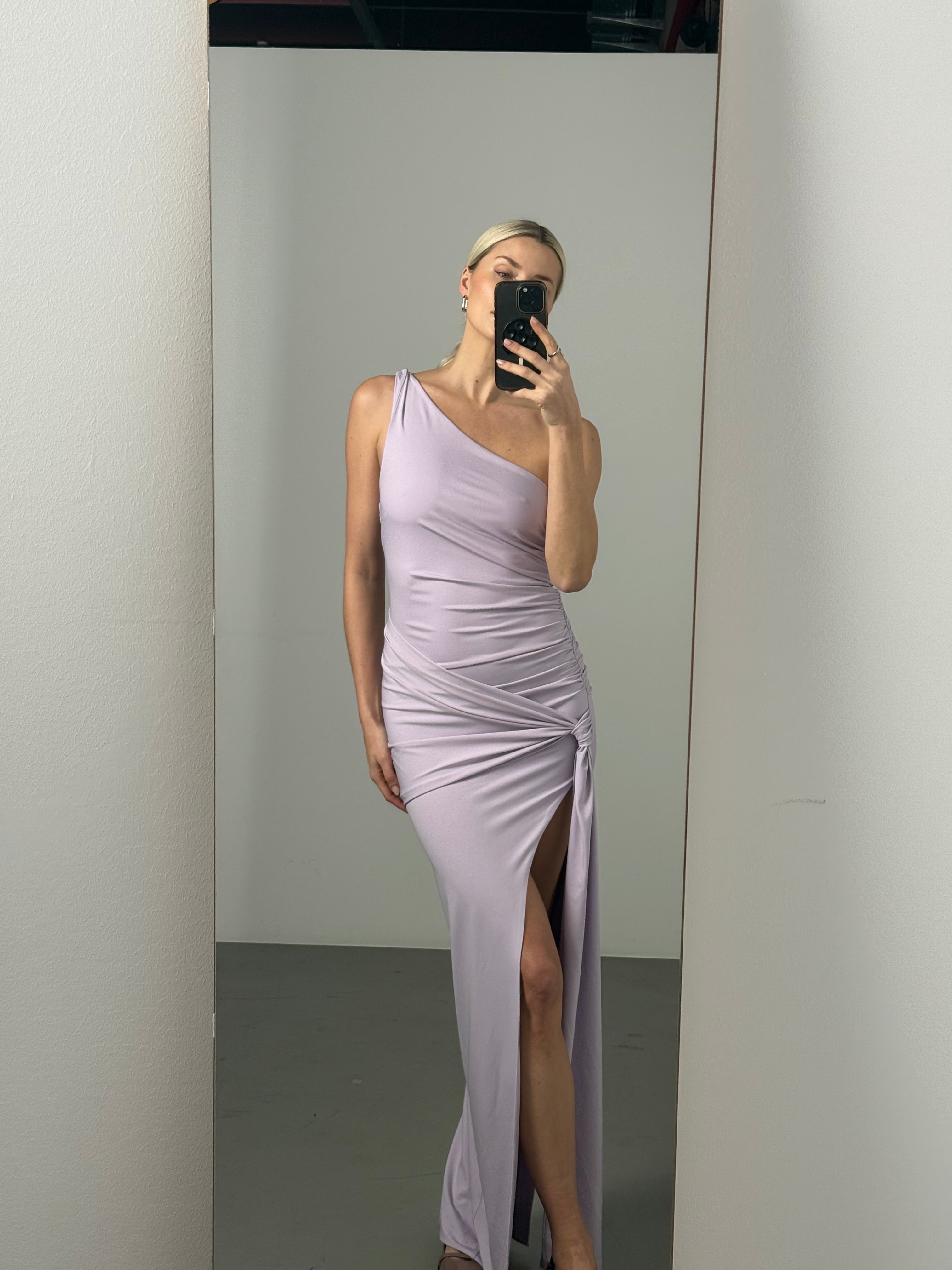 Diese Person trägt das Claire Dress von LeGer by Lena Gercke, ein hellviolettes, schulterfreies Kleid mit oberschenkelhohem Schlitz, und macht ein Spiegel-Selfie in einem minimalistischen, weiß getünchten Raum.