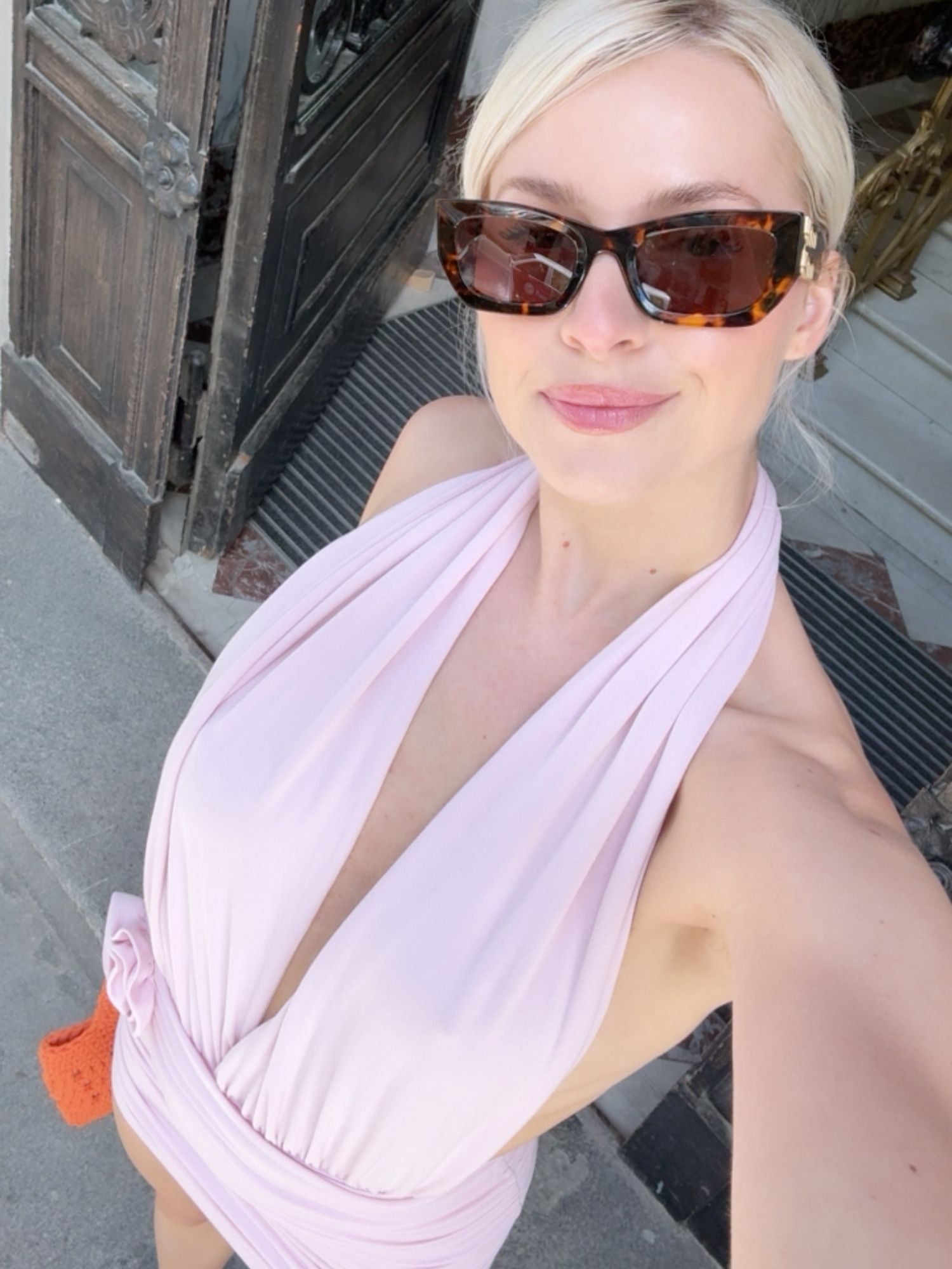 Eine Frau mit blonden Haaren, die zu einem tief sitzenden Dutt zusammengebunden sind, trägt eine große Sonnenbrille in Schildpattoptik und das zartrosa „Rosie“-Kleid von LeGer by Lena Gercke. Sie macht draußen in der Nähe einer Holztür ein Selfie, lächelt dabei leicht und hält eine orangefarbene, strukturierte Tasche in der Hand.