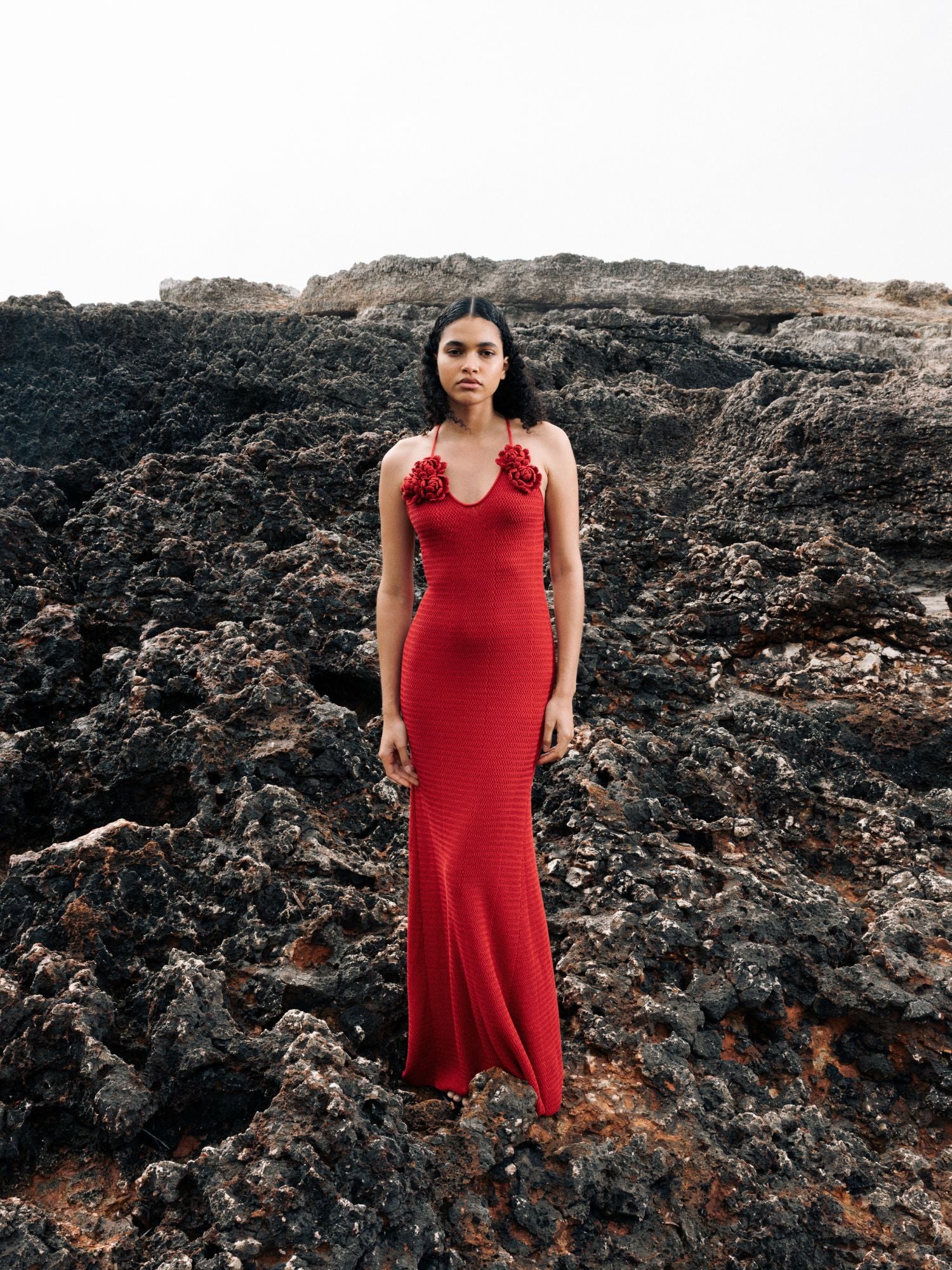 Eine Frau steht auf dunklem, felsigem Gelände und posiert in dem Kleid „Lia“ von LeGer by Lena Gercke – einem roten Maxikleid mit 3D-Blumenrosetten im Brustbereich und einer Meerjungfrauen-Silhouette – vor dramatischen Vulkangesteinen unter einem bewölkten Himmel.