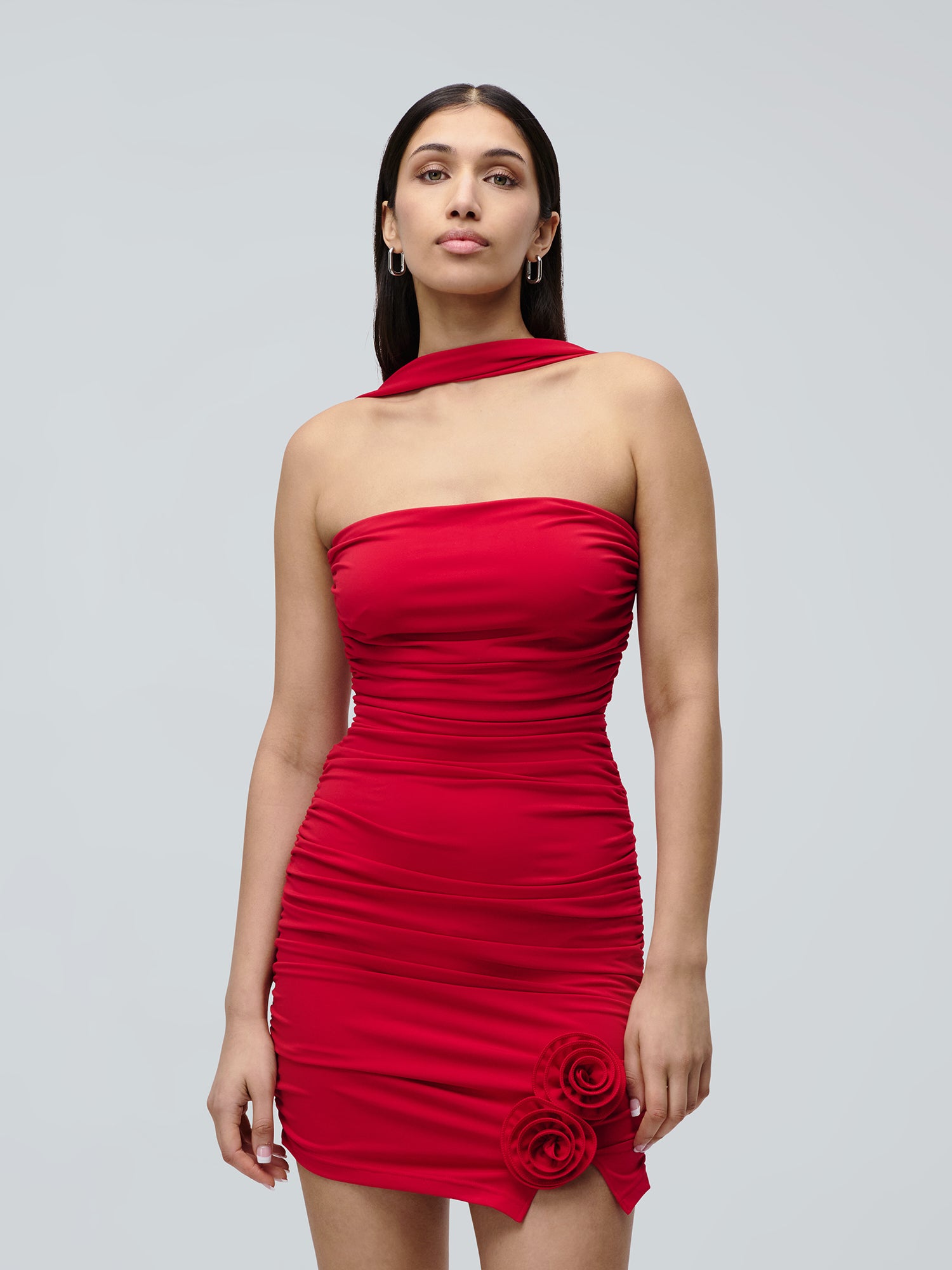 Eine Frau im Kleid „Eike“ von LeGer by Lena Gercke posiert vor einem hellgrauen Hintergrund. Sie trägt das trägerlose, geraffte rote Minikleid mit zwei Stoffrosen und einem einzigartigen Ausschnitt. Sie hat glattes dunkles Haar, Creolen und einen neutralen Gesichtsausdruck.