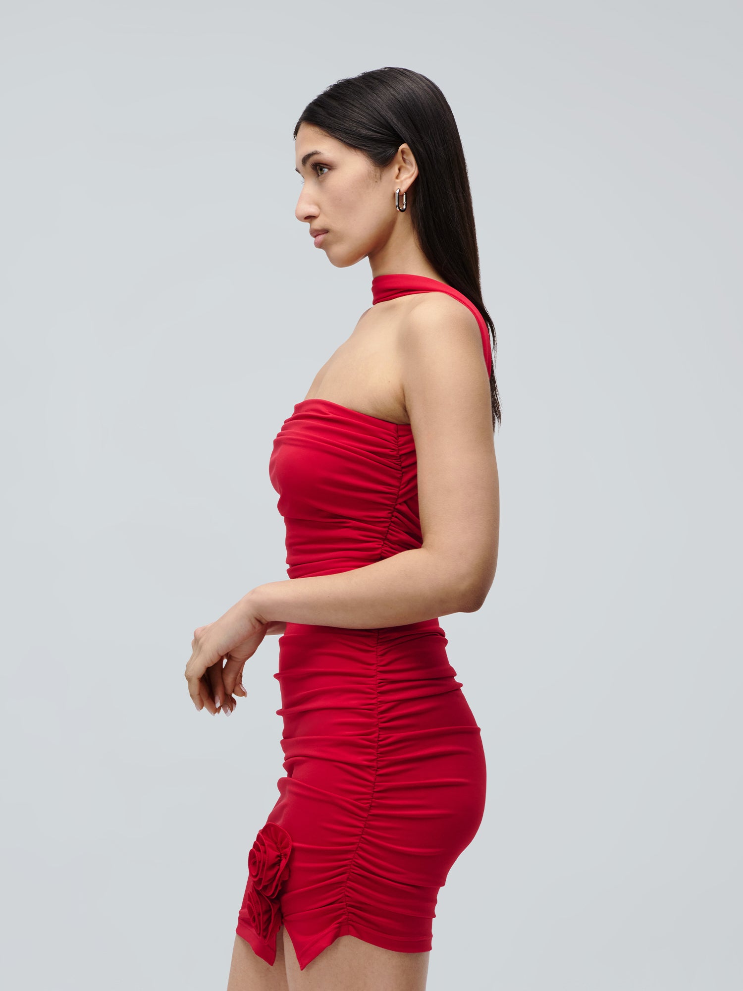 Eine Frau im Kleid „Eike“ von LeGer by Lena Gercke – ein eng anliegendes, ärmelloses rotes Minikleid mit Rüschen und Rosenverzierung – steht im Profil vor einem schlichten hellgrauen Hintergrund. Sie hat langes, glattes, dunkles Haar und trägt Creolen.