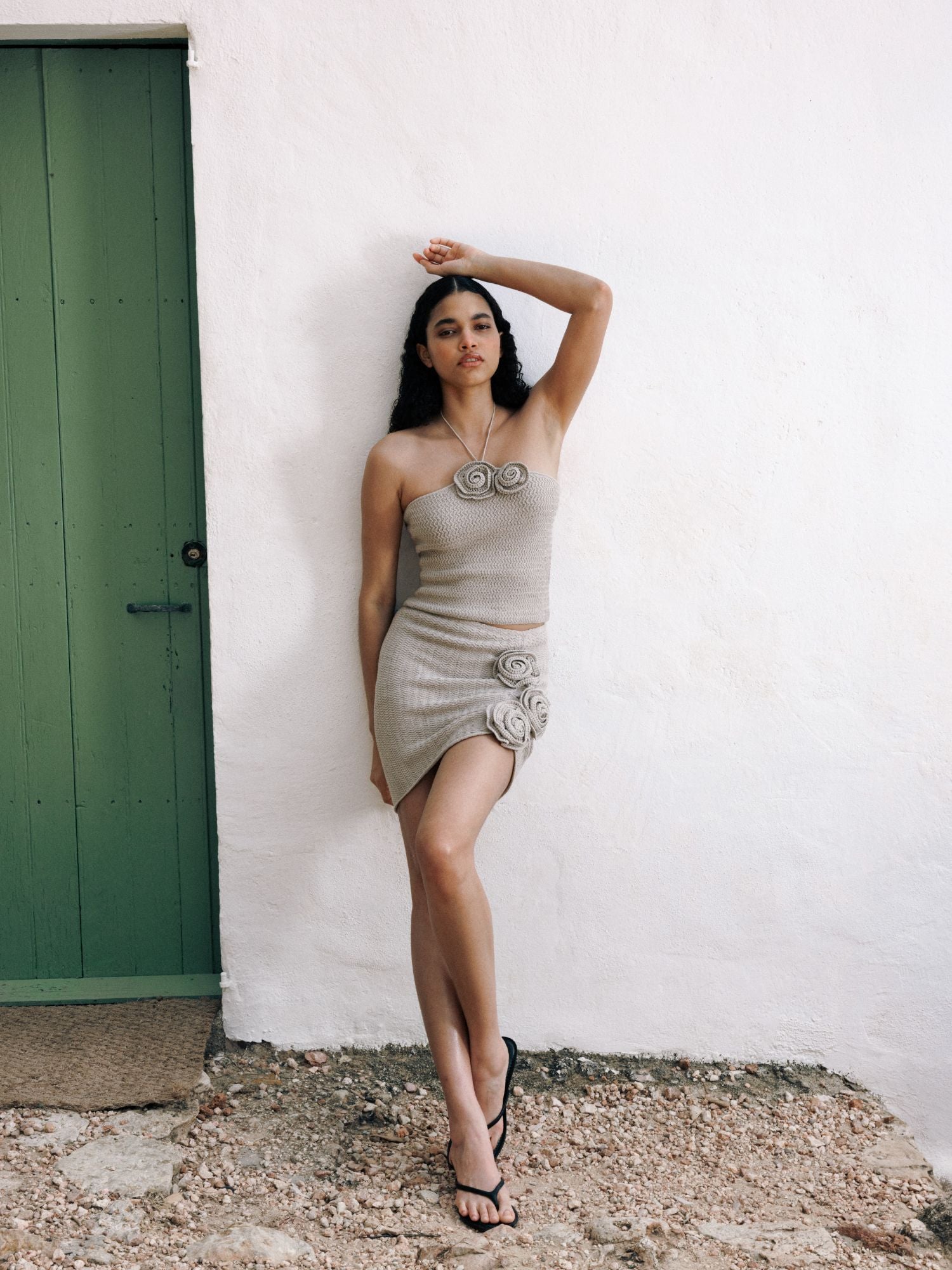 Eine Frau, die den beigefarbenen, hoch taillierten Rock Lia von LeGer by Lena Gercke mit Blumendetails trägt, steht selbstbewusst vor einer weißen Wand in der Nähe einer grünen Tür, posiert mit erhobenem Arm und kombiniert den Look mit schwarzen Sandalen.