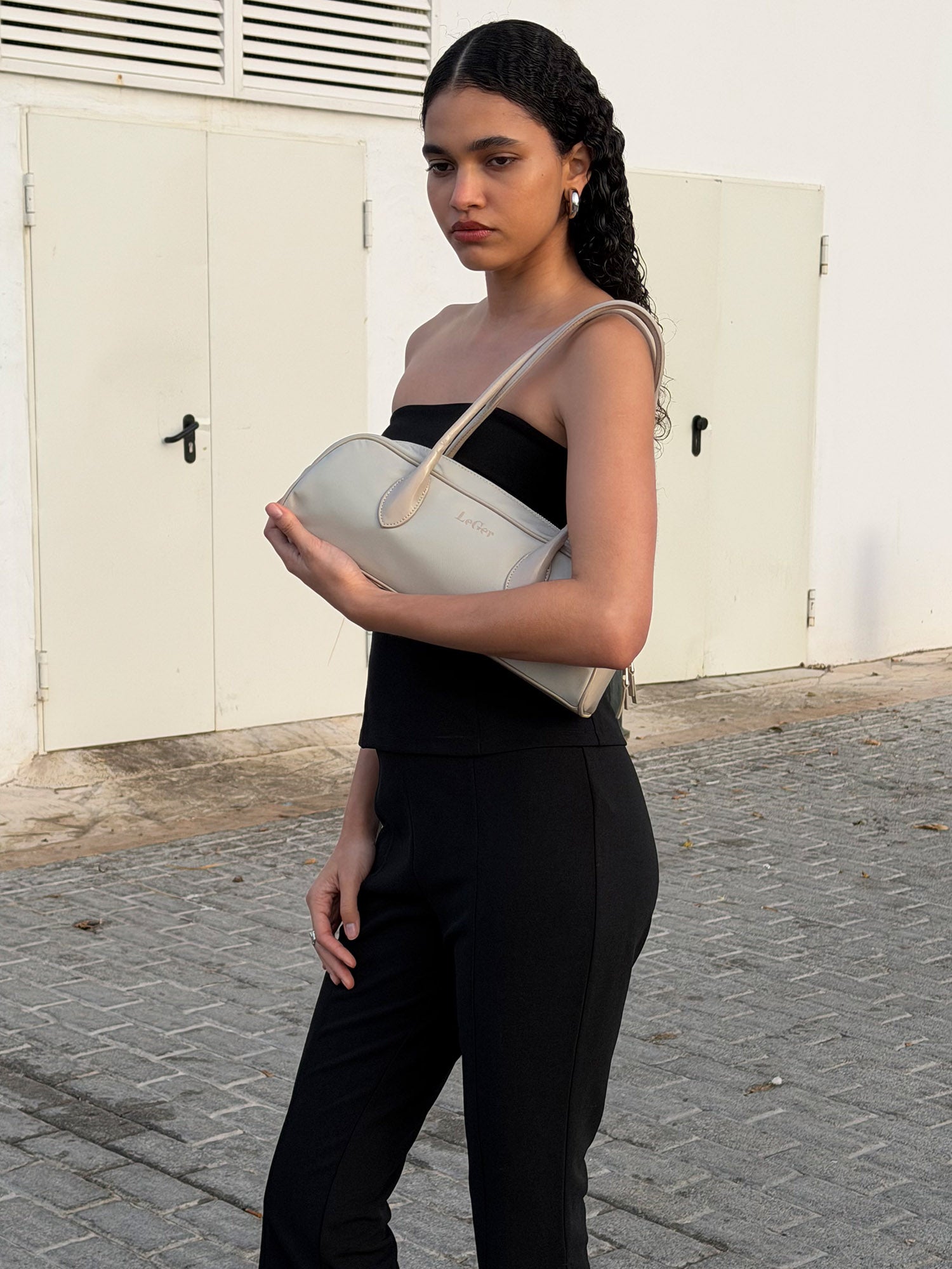 Eine Frau mit lockigem Haar steht im Freien und trägt das Dunja Set von LeGer by Lena Gercke - bestehend aus einem schwarzen trägerlosen Top und schwarzen Capri-Hosen - mit einer beigen Handtasche in der Hand, hinter ihr weiße Türen und Backsteinpflaster.