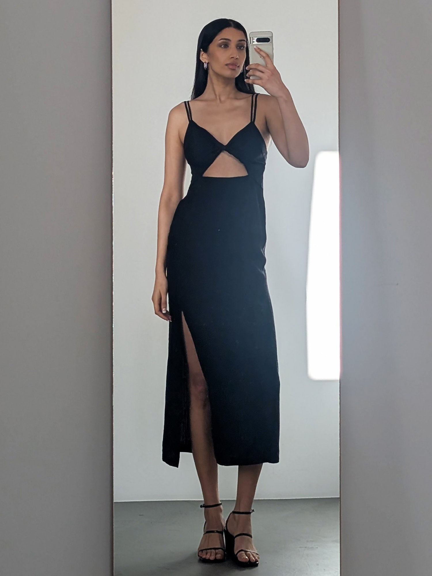 Eine Frau posiert für ein Selfie im Spiegel in dem Cay Dress von LeGer by Lena Gercke - einem schwarzen, eleganten Damen-Kleid mit dünnen Trägern, einem Ausschnitt in der Taille und einem oberschenkelhohen Schlitz -, das mit hochhackigen Sandalen in einer minimalen, gut beleuchteten Umgebung gestylt ist.