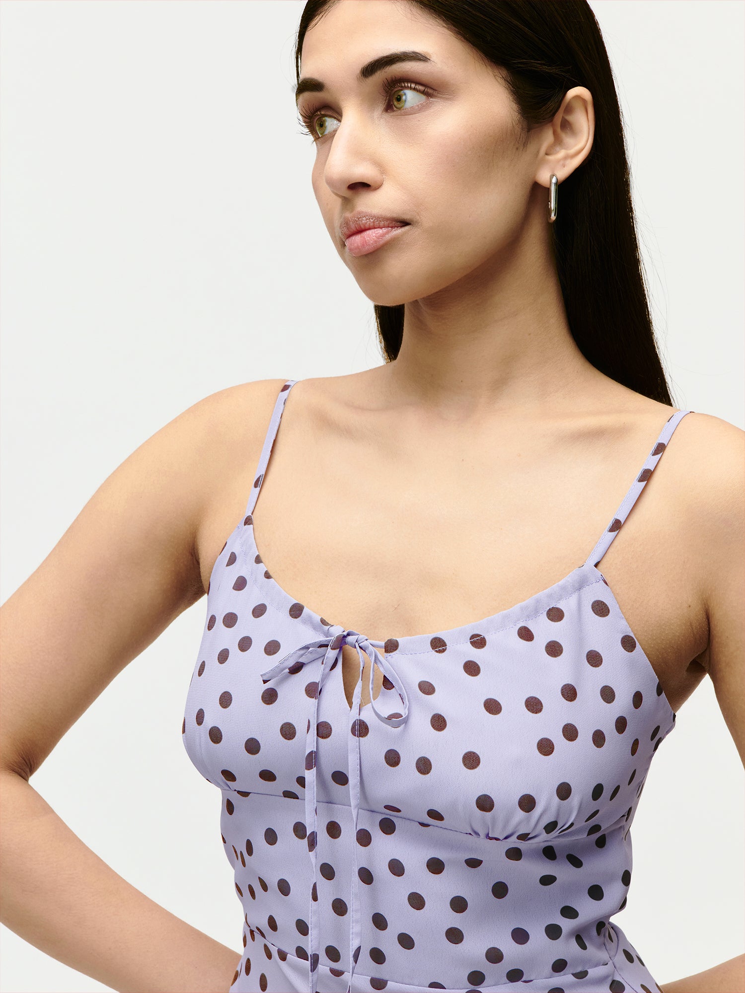 Eine Frau mit langen dunklen Haaren trägt das Fleur Dress von LeGer by Lena Gercke - ein lavendelfarbenes, ärmelloses Kleid mit schwarzen Tupfen und einem Bindeband auf der Vorderseite - und zeigt damit einen selbstbewussten Sommerlook vor einem schlichten, hellen Hintergrund.
