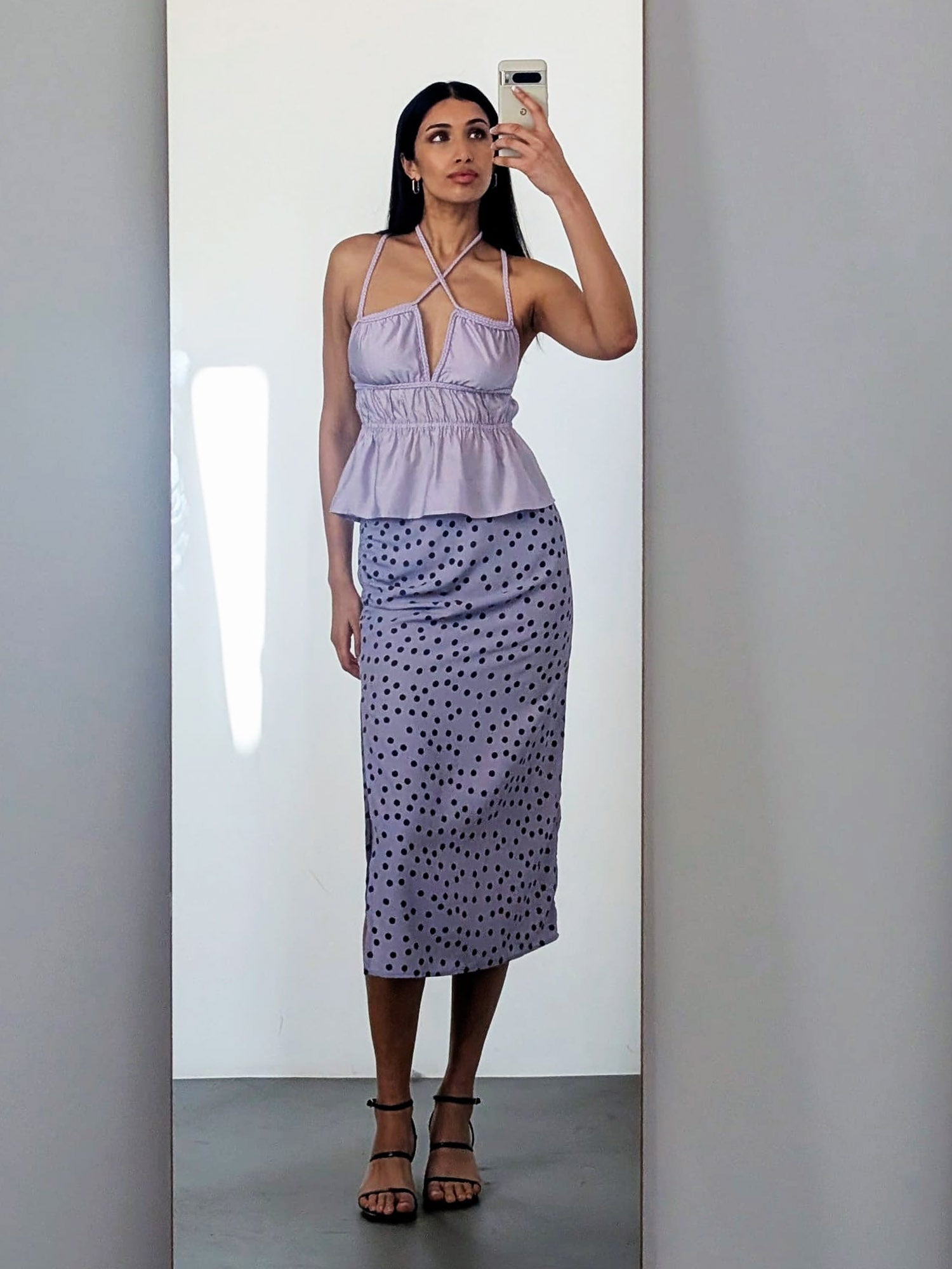 Eine Frau mit langen dunklen Haaren macht ein Selfie im Fabrice Top von LeGer by Lena Gercke und einem passenden lilafarbenen gepunkteten Midirock, der mit schwarzen Riemchensandalen für einen schicken, modernen Look sorgt.