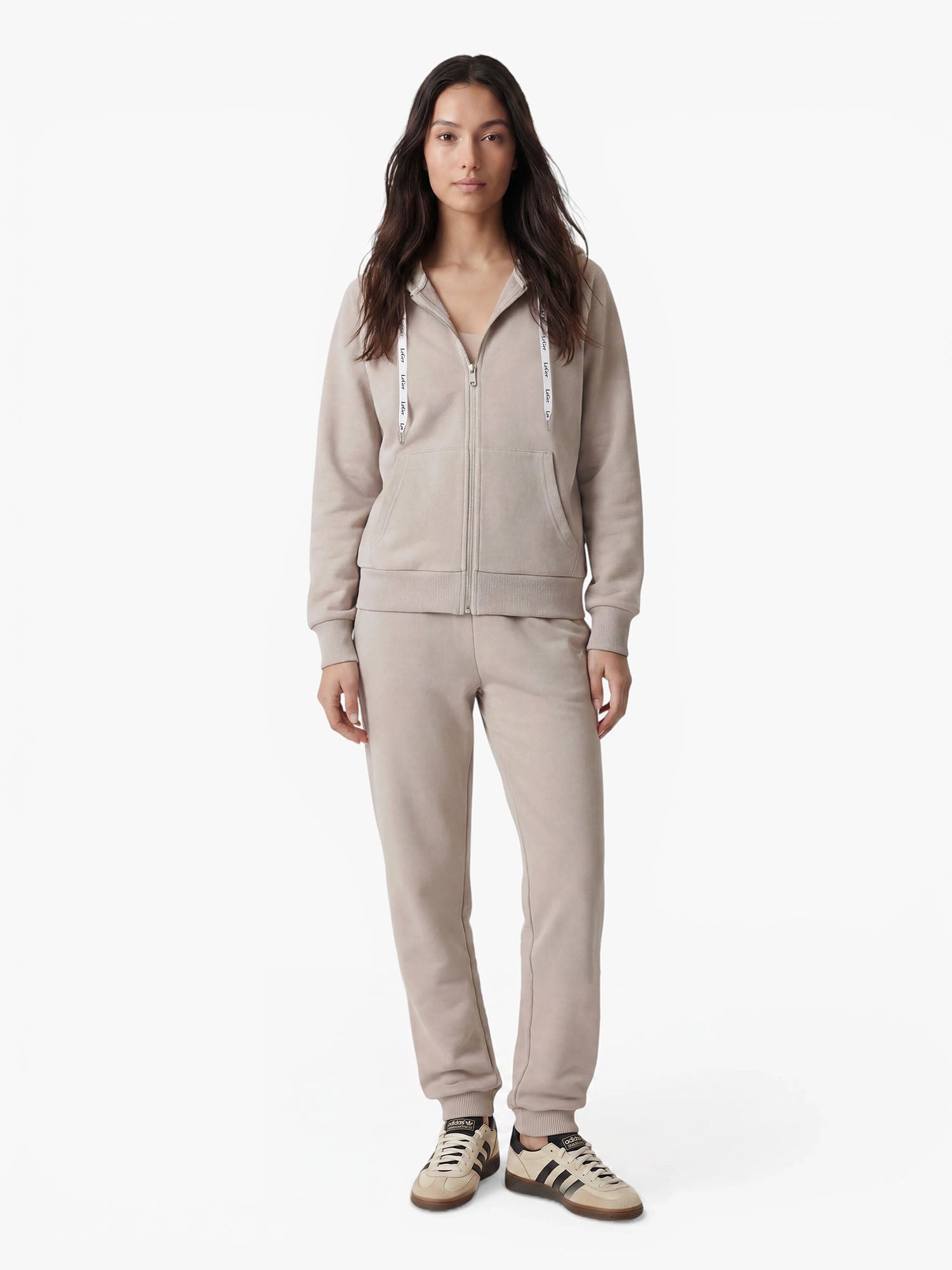 Eine Frau steht mit dem Blick nach vorne und strahlt Komfort aus, während sie den hellbeigen „Phoenix“-Hoodie von LeGer by Lena Gercke mit passender Jogginghose und beigen Sneakers mit schwarzen Streifen trägt – perfekt für sportliche Aktivitäten – vor einem schlichten weißen Hintergrund.