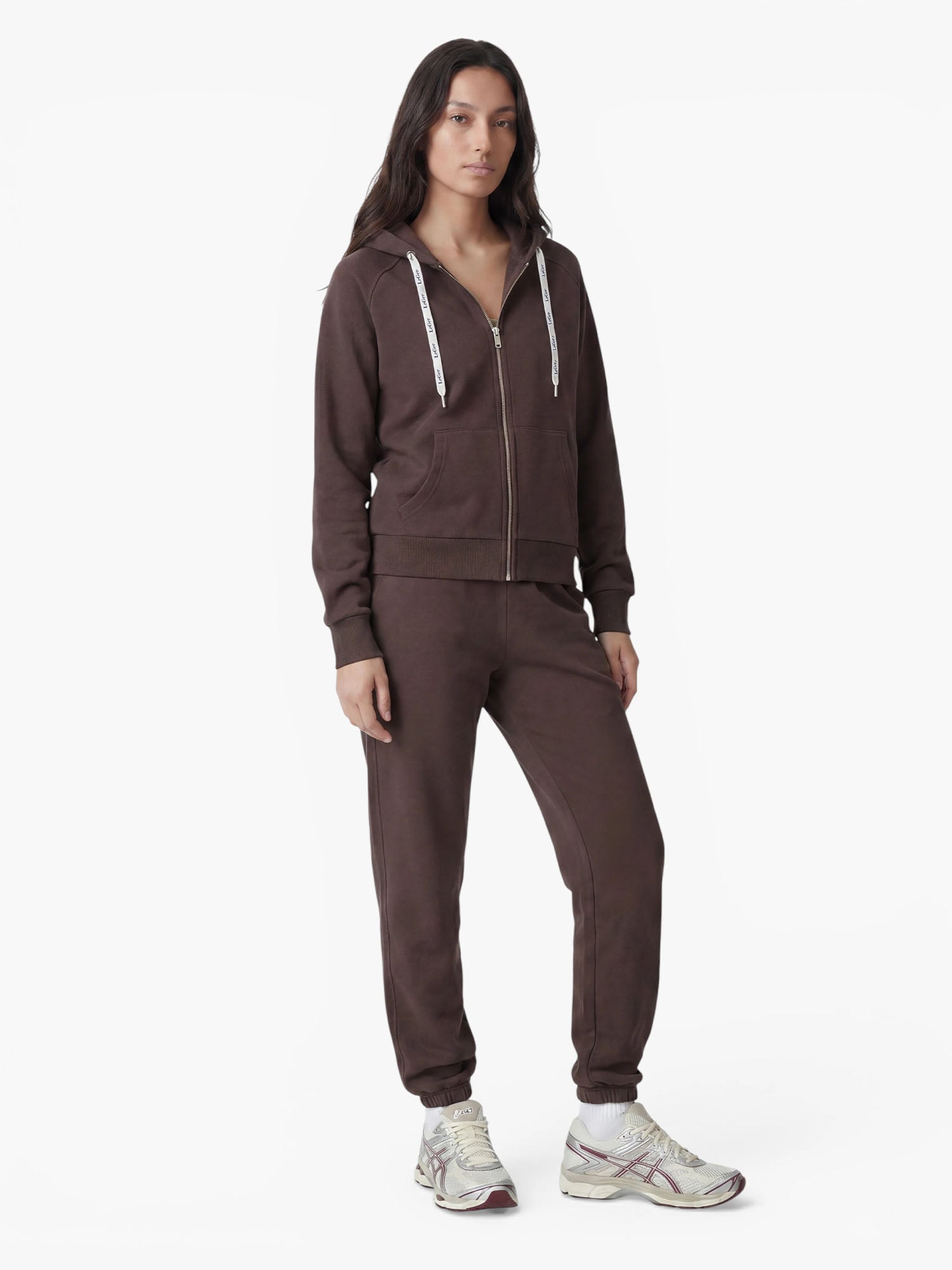 Eine Person steht vor einem schlichten weißen Hintergrund und trägt den „Phoenix“-Hoodie von LeGer by Lena Gercke in Dunkelbraun mit strapazierfähigem Design, dazu eine passende Jogginghose und weiße Sneaker.