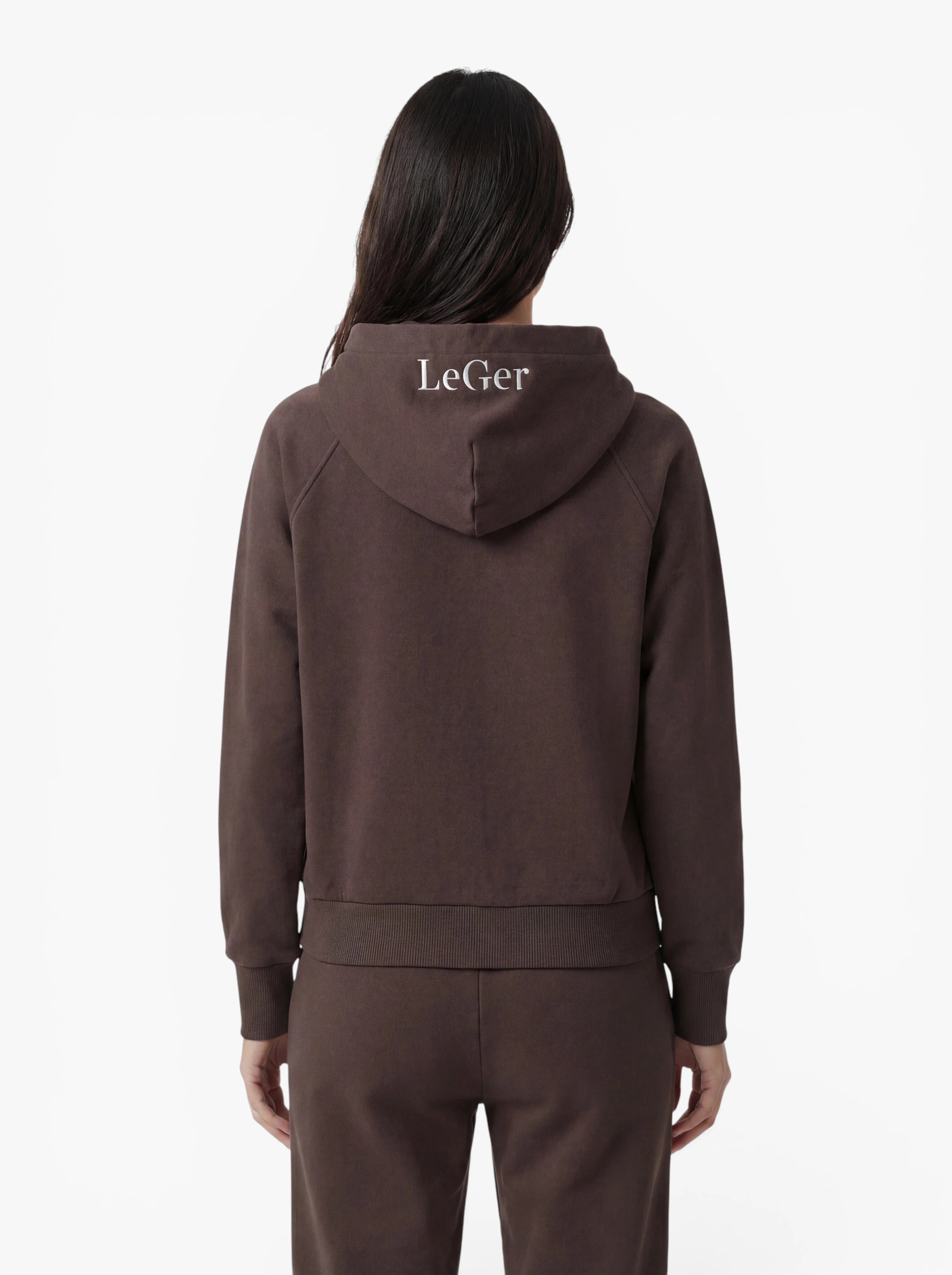 Eine Person mit langen, dunklen Haaren trägt den braunen „Phoenix“-Hoodie von LeGer by Lena Gercke, der sich durch einen sportlichen Schnitt auszeichnet, sowie eine dazu passende Hose – hier von hinten gesehen. Auf der Kapuze ist das weiße „LeGer“-Logo aufgedruckt, und der schlichte weiße Hintergrund unterstreicht das strapazierfähige Design.