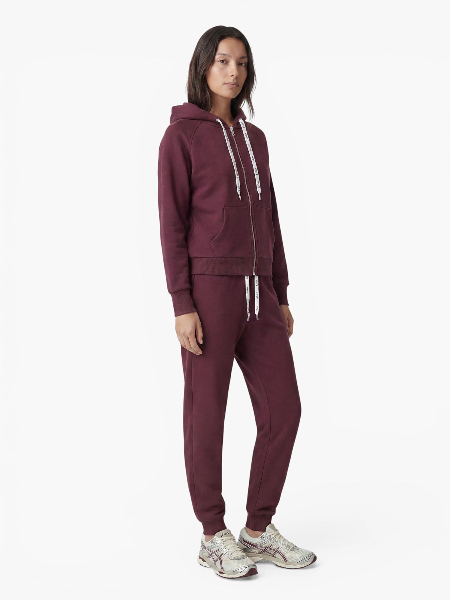 Eine Person steht mit dem Gesicht nach vorne und trägt einen burgunderroten „Phoenix“-Kapuzenpullover von LeGer by Lena Gercke sowie eine dazu passende Jogginghose. Sie hat mittellanges dunkles Haar und trägt dazu weiße und silberne Sneaker. Der sportliche Kapuzenpullover aus weicher Baumwollmischung sorgt für besonderen Tragekomfort. Der Hintergrund ist schlicht weiß.