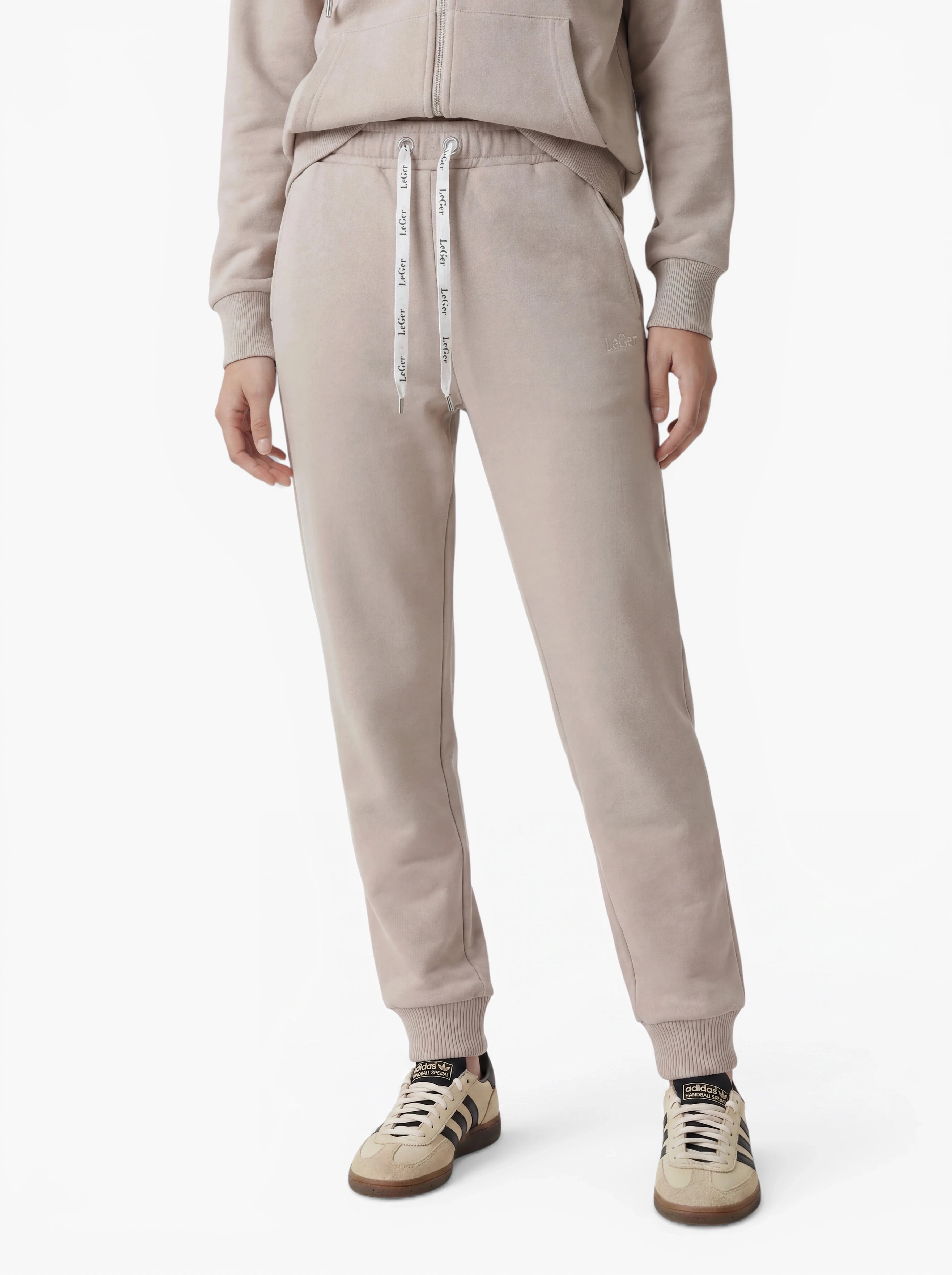 Vor einem weißen Hintergrund steht eine Person in einer hellbeigen „Nela“-Hose von LeGer by Lena Gercke mit Kordelzug in der Taille, dazu ein passender Hoodie und cremefarbene Sneaker mit schwarzen Akzenten – ein Outfit, das eine moderne Silhouette mit hochwertigen Materialien vereint.
