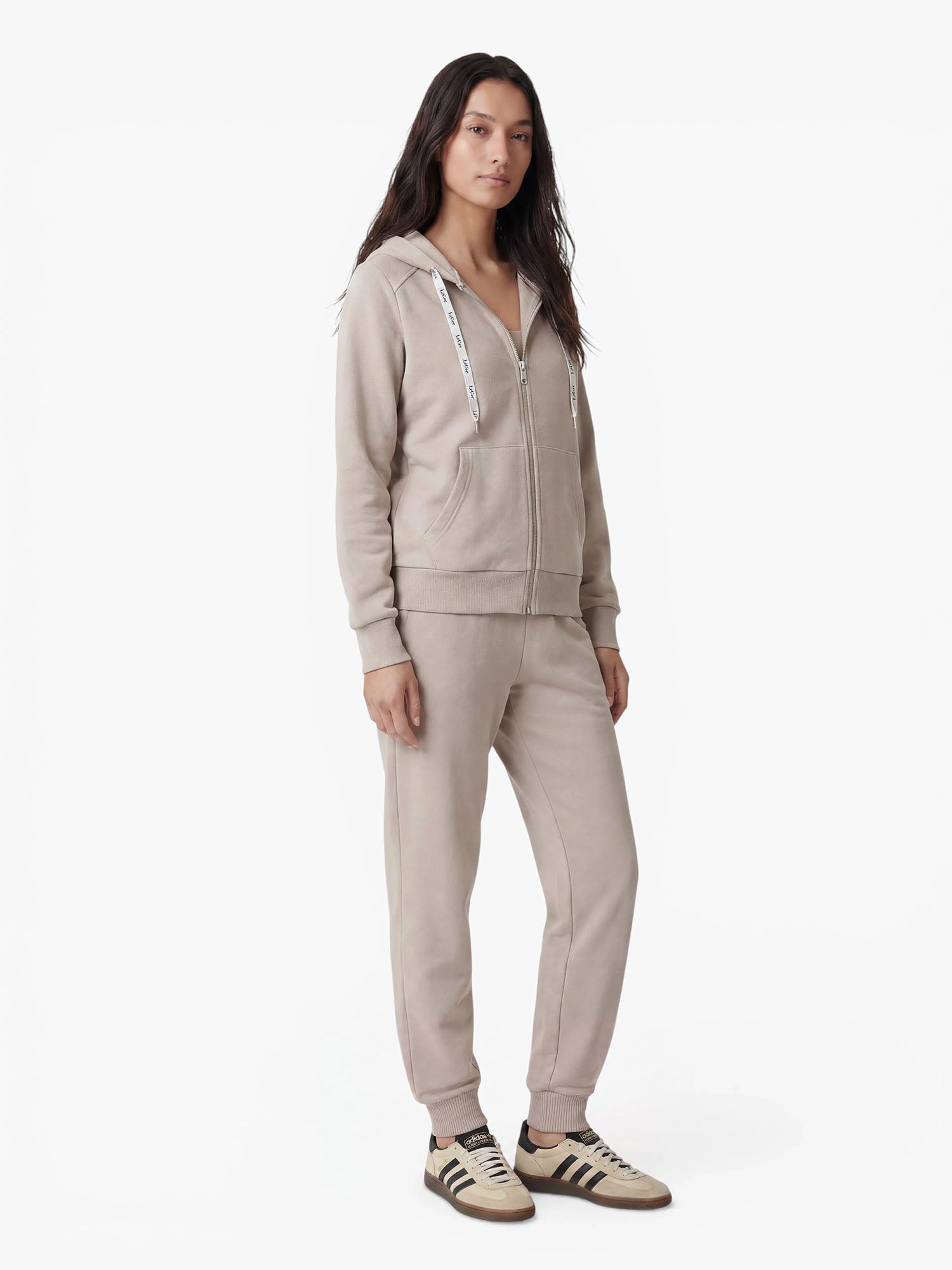 Eine Frau steht mit dem Blick nach vorne vor einem schlichten weißen Hintergrund. Sie trägt einen taupefarbenen Kapuzenpullover mit Reißverschluss, dazu die „Nela“-Hose von LeGer by Lena Gercke, die sich durch eine moderne Silhouette auszeichnet, sowie beige-schwarze Sneaker.